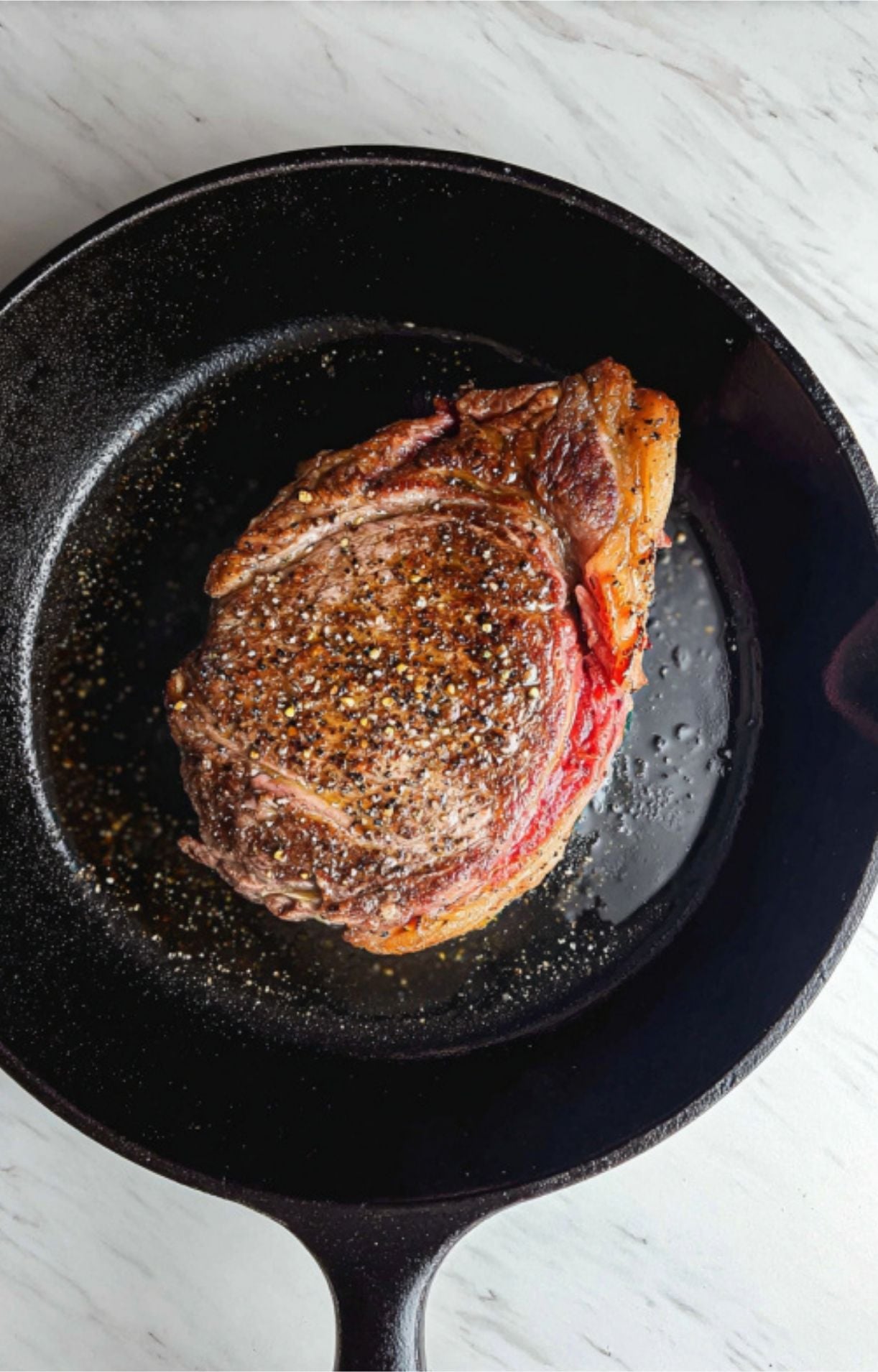 A rib eye steak cooking in a cast-iron skillet, showing a beautifully developed golden-brown crust and glistening rendered fat.