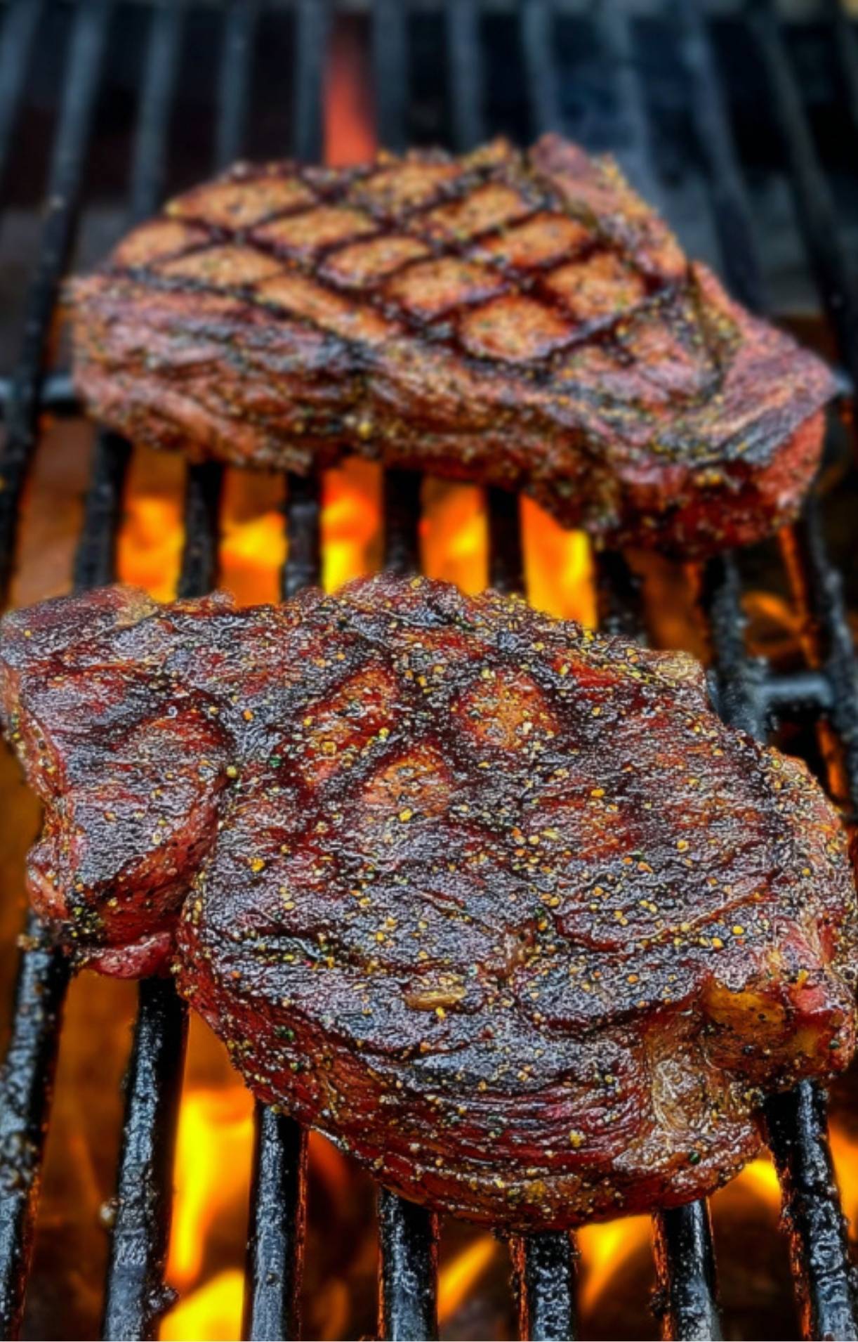 Two seasoned steaks cooking over high heat on grill grates with visible orange flames and diamond-patterned char marks.