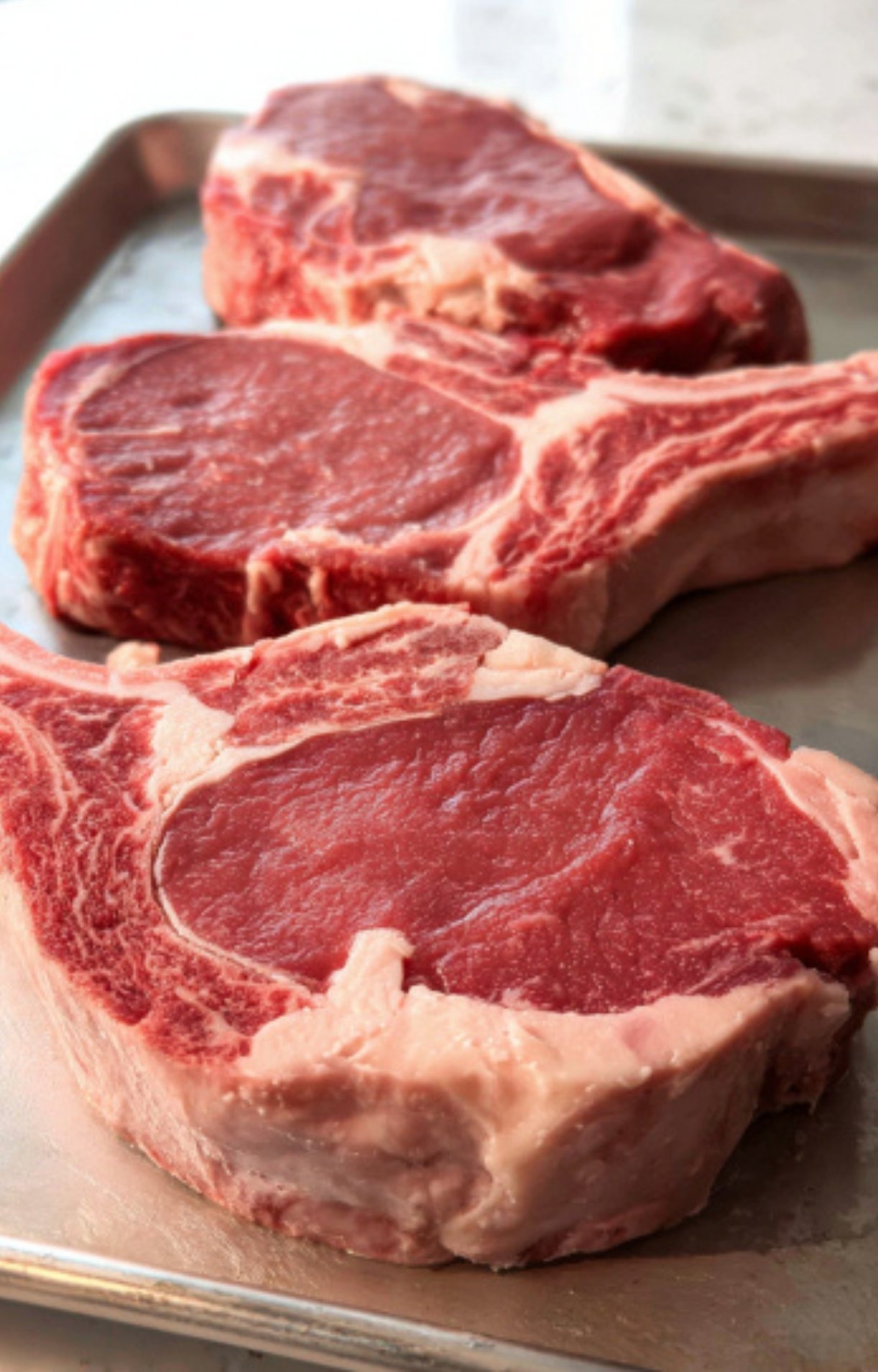 Three thick, raw bone-in rib eye steaks arranged on a silver baking sheet, showing the rich marbling and deep red color of the beef.