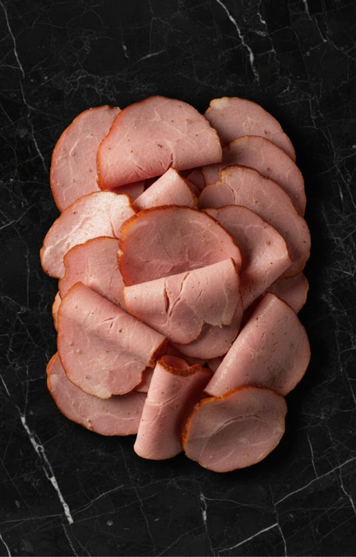 A top-down view of round, thin slices of smoked ham arranged on a black marble surface before packaging.