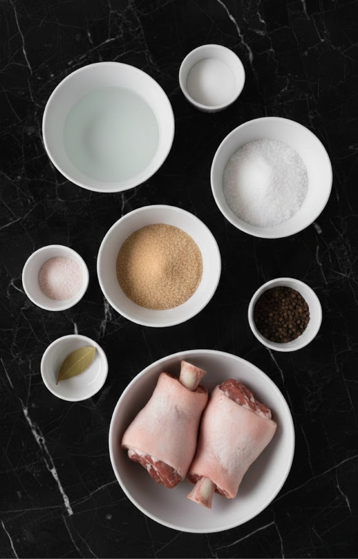An overhead shot of white bowls containing water, salt, brown sugar, curing salt, peppercorns, a bay leaf, and raw pork hocks.