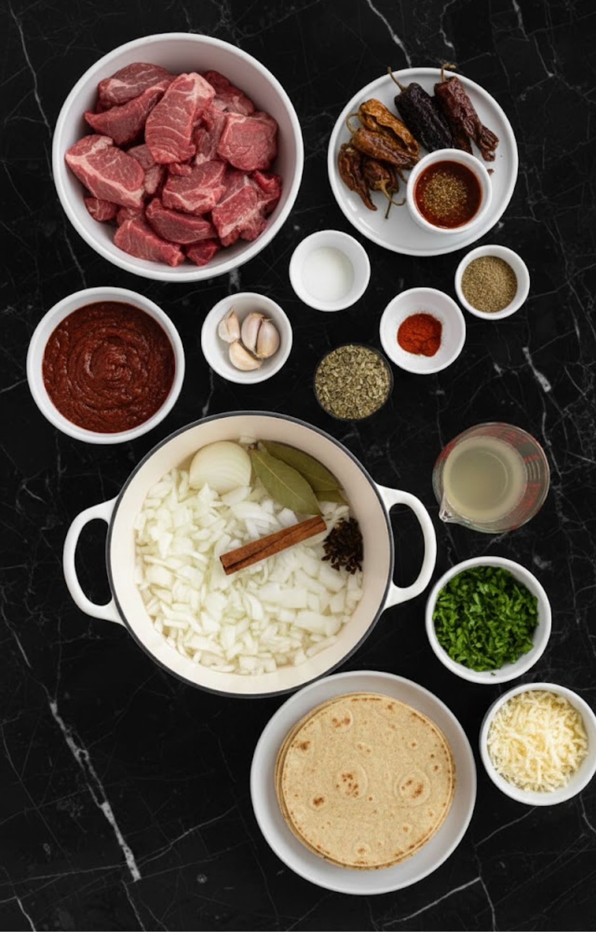 An overhead shot of ingredients on a black marble surface, including cubed beef, dried chiles, chili paste, corn tortillas, shredded cheese, and a pot with onions and whole spices.