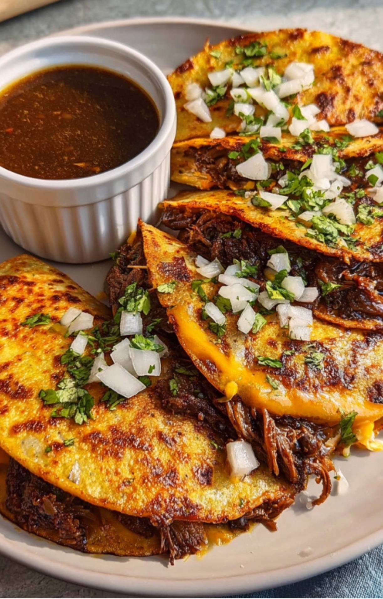 A close-up view of three crispy quesa birria tacos topped with fresh diced white onions and chopped cilantro, served with a side of rich dark consommé in a white ramekin.