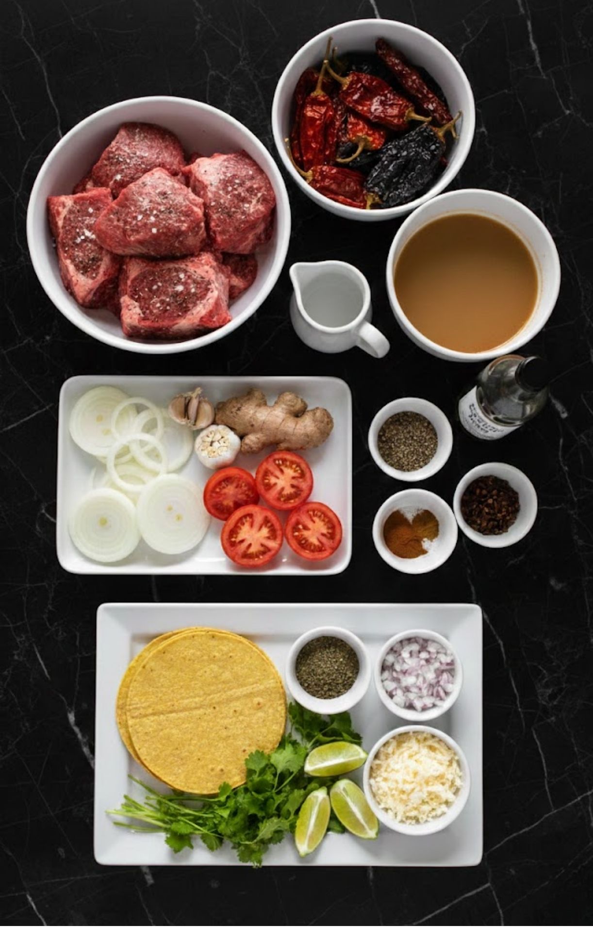 A top-down view of organized ingredients including beef chunks, dried chilies, beef broth, sliced onions, tomatoes, ginger, garlic, and taco garnishes like cilantro and lime.