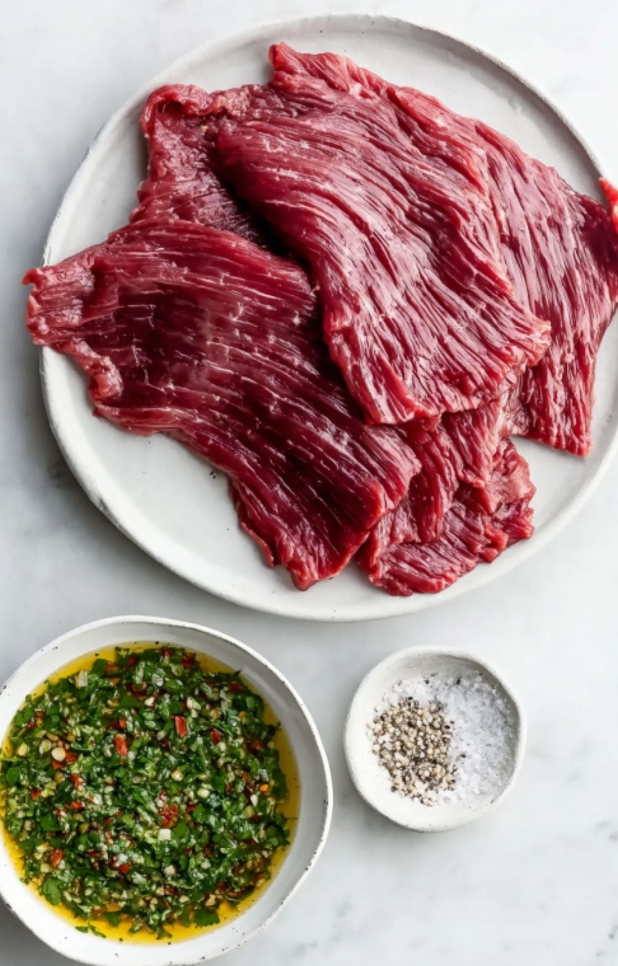 Fresh raw skirt steak laid out on a plate next to bowls of homemade chimichurri sauce, cracked pepper, and sea salt, ready for grilling.