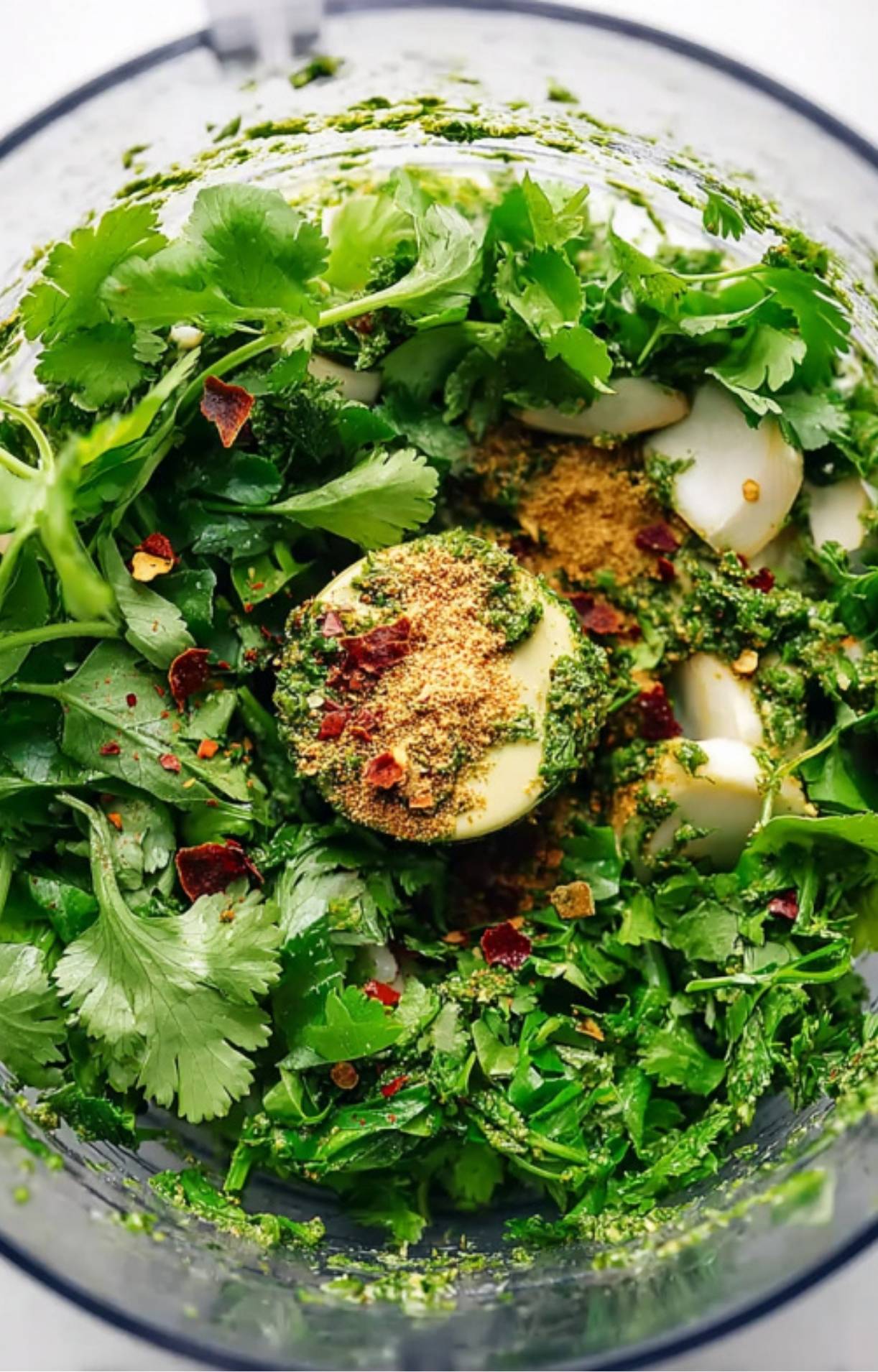 Food processor filled with cilantro, parsley, garlic, spices, and red pepper flakes. Ingredients for making the best avocado chimichurri sauce.