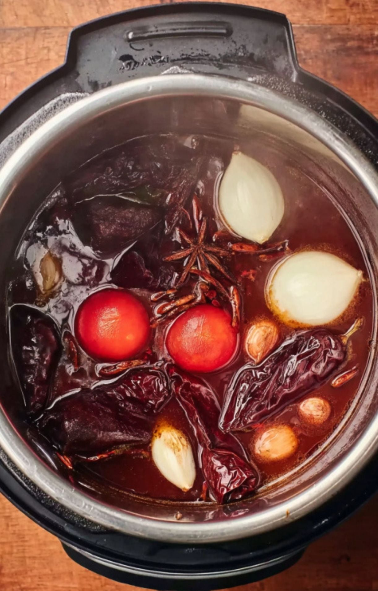An Instant Pot filled with beef broth, dried red chilies, whole tomatoes, onions, garlic cloves, and star anise being prepared for pressure cooking.