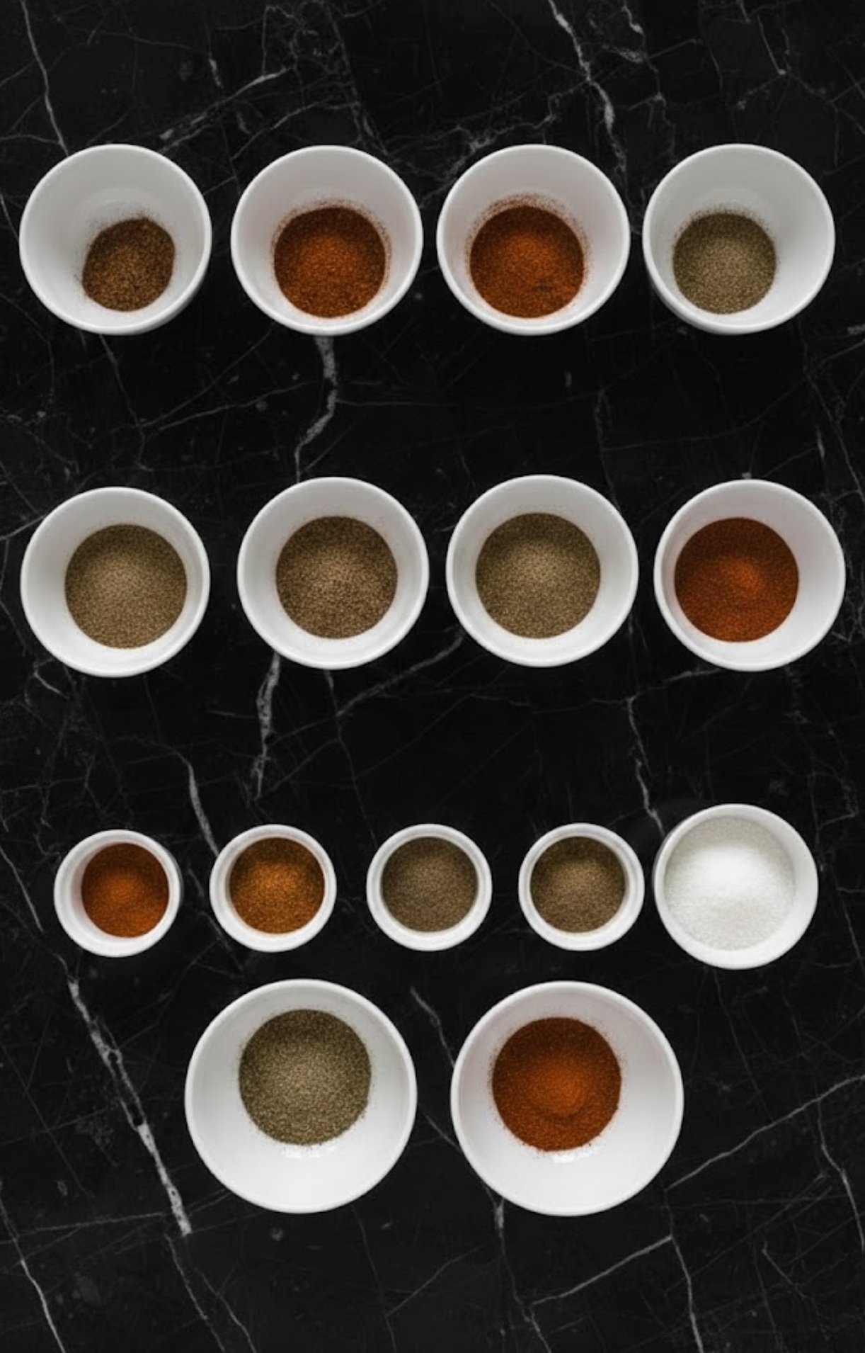 A top-down view of whole dried ancho and guajillo chilies in a white bowl, accompanied by small plates of cinnamon sticks, black peppercorns, cloves, ginger powder, cumin, dried oregano, and salt.