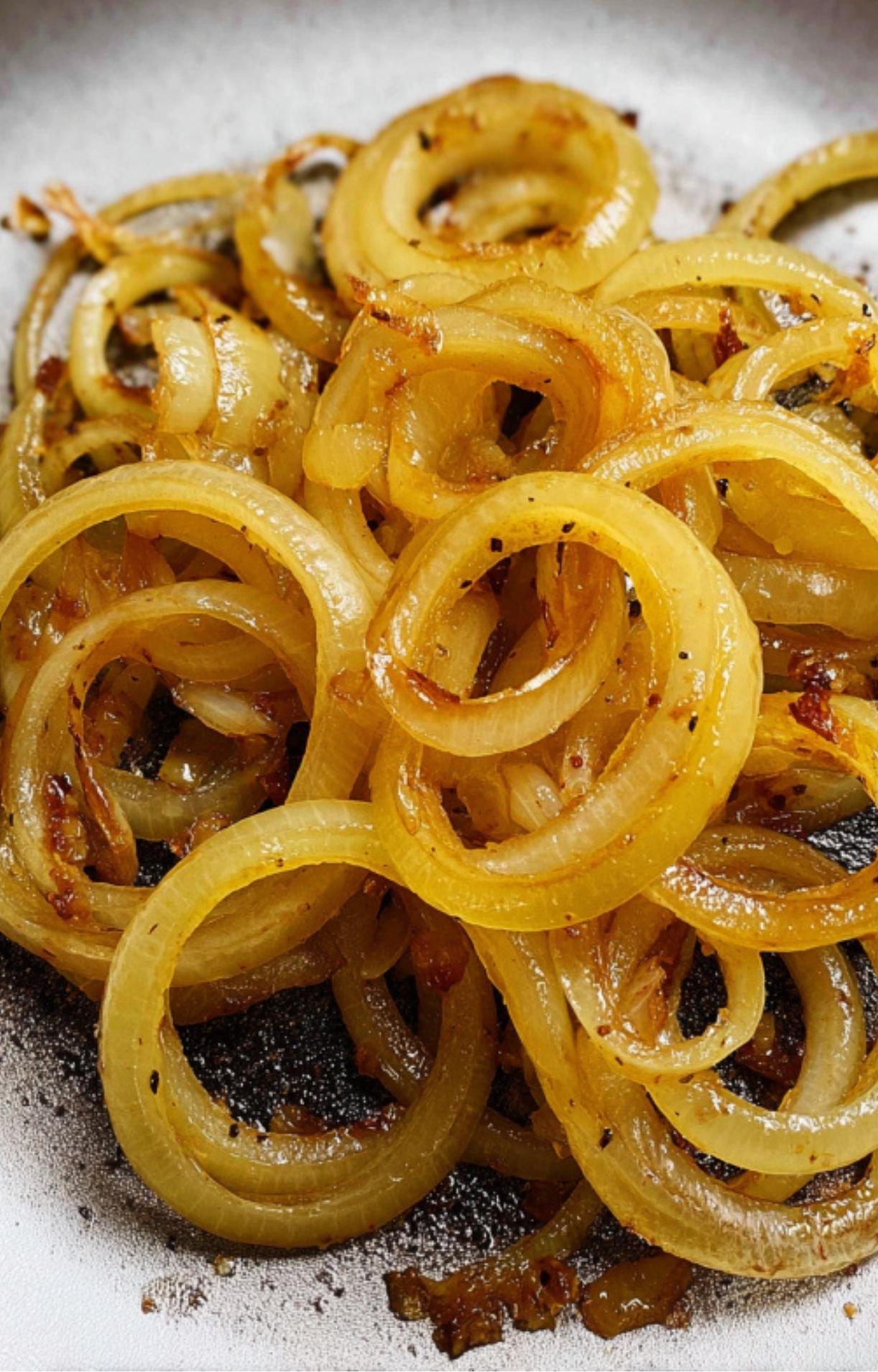 A close-up of golden-brown sautéed onion rings glistening with oil and spices in a pan.