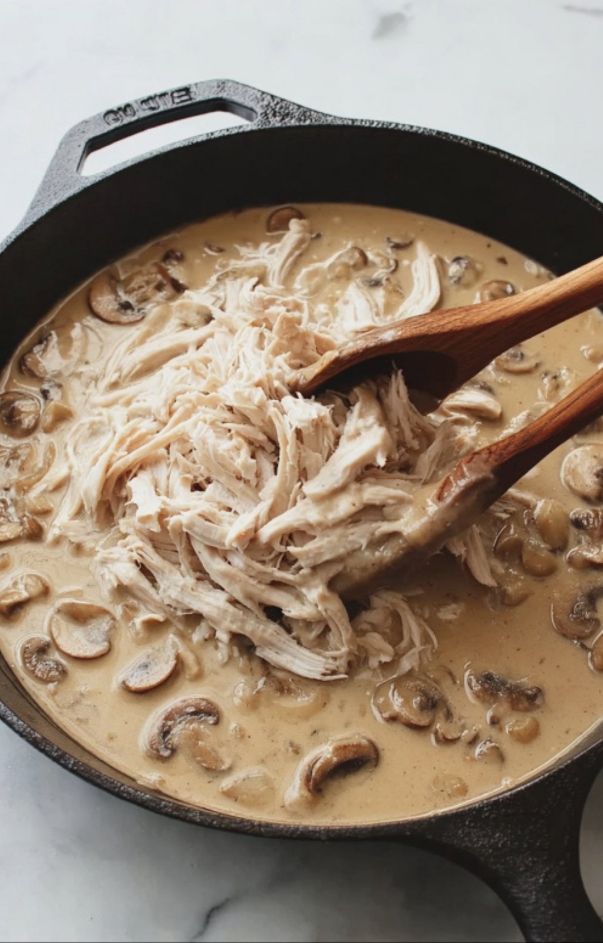 Two wooden spoons folding a large pile of shredded leftover turkey into a creamy mushroom sauce inside a skillet.