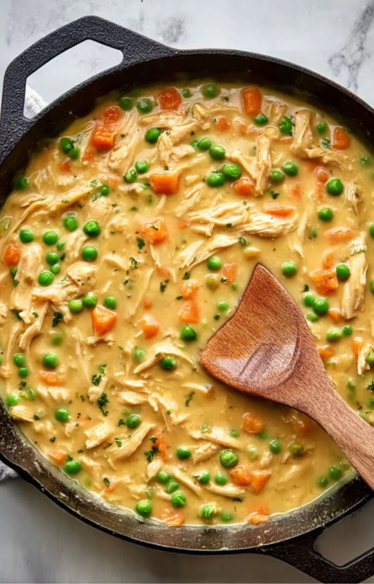 A wooden spoon stirring shredded turkey and bright green peas into a thick, creamy vegetable gravy for Gluten Free Turkey Pot Pie.