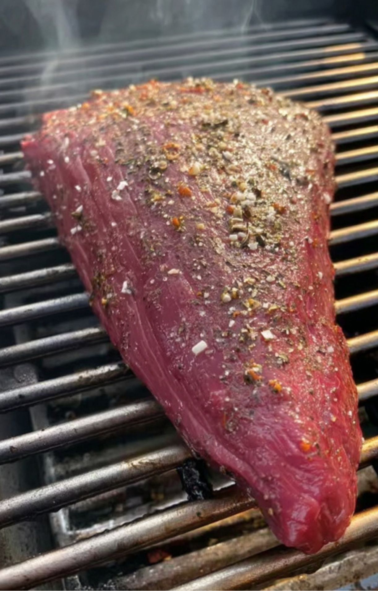 Uncooked tri tip steak sprinkled with salt and pepper, placed on grill grates with smoke rising.