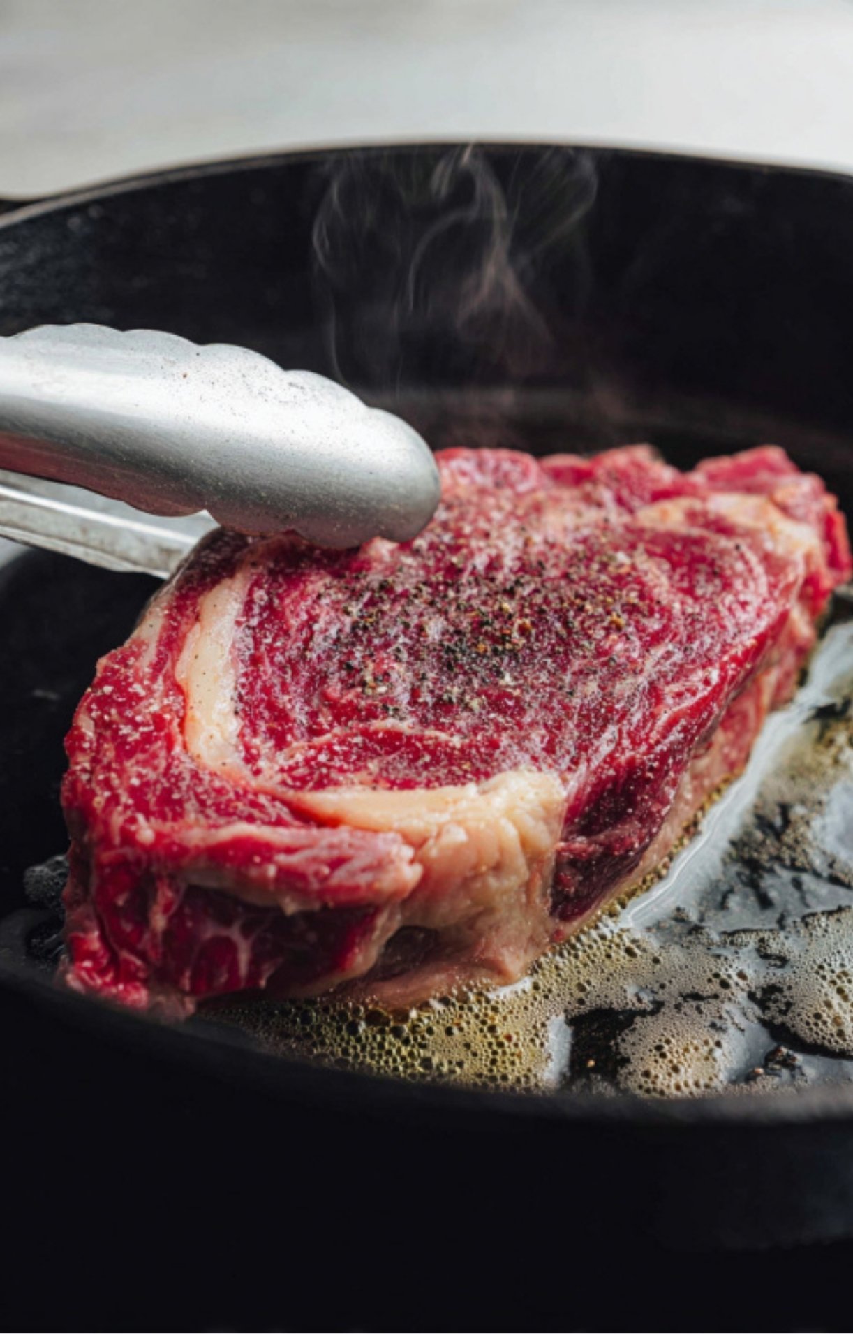 Using metal tongs to carefully place a thick-cut, seasoned rib eye steak into a sizzling cast-iron pan with hot oil and rising steam.