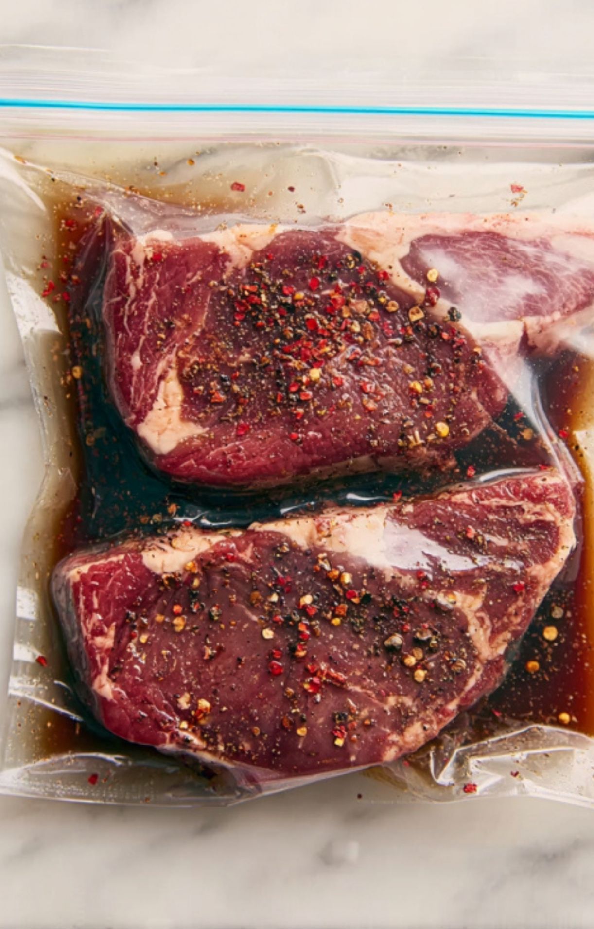 Raw rib eye steaks soaking in a dark savory marinade with peppercorns inside a sealed plastic bag.