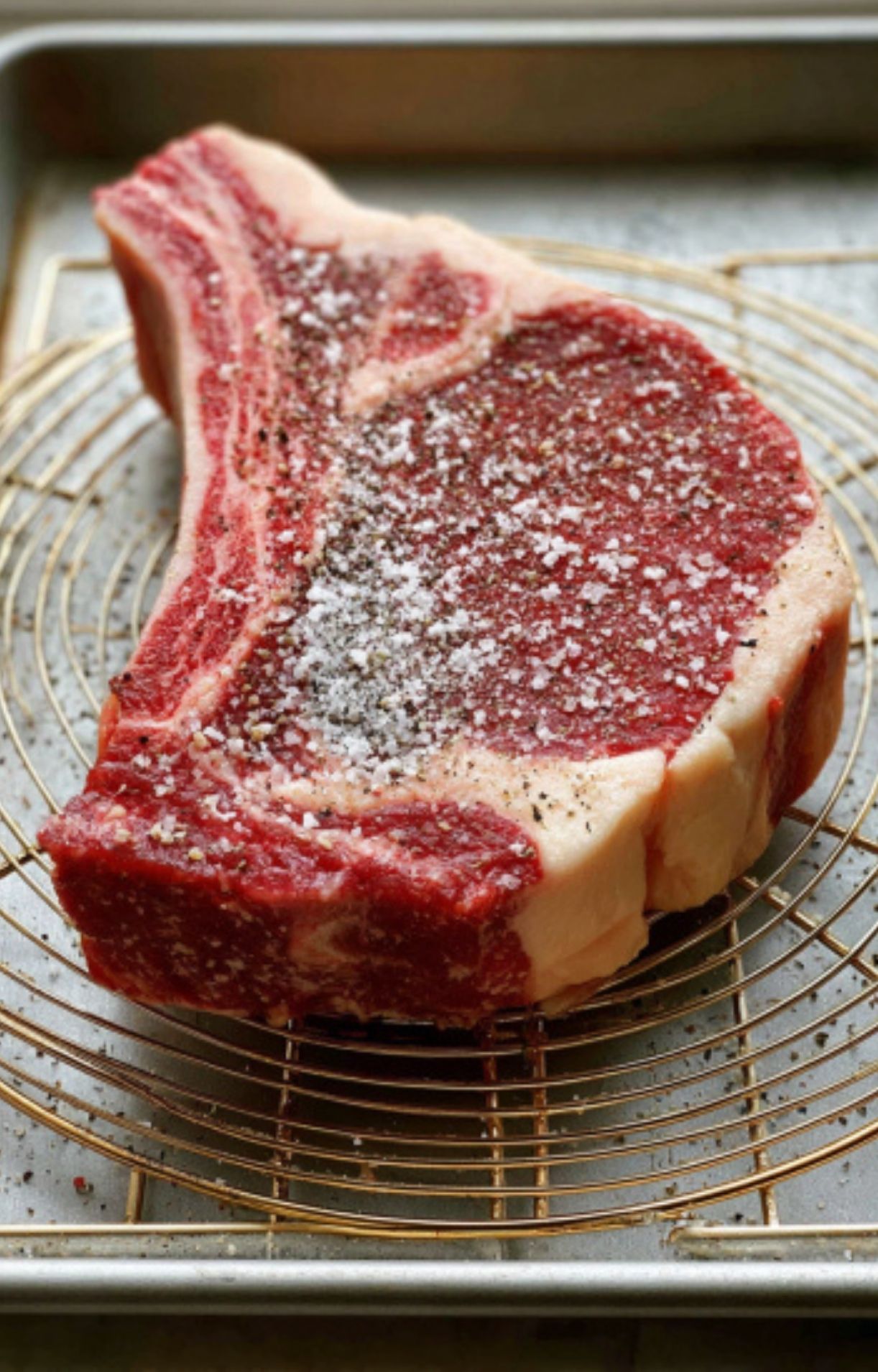 A thick bone-in rib eye steak resting on a wire rack, generously coated with coarse salt and cracked black pepper.