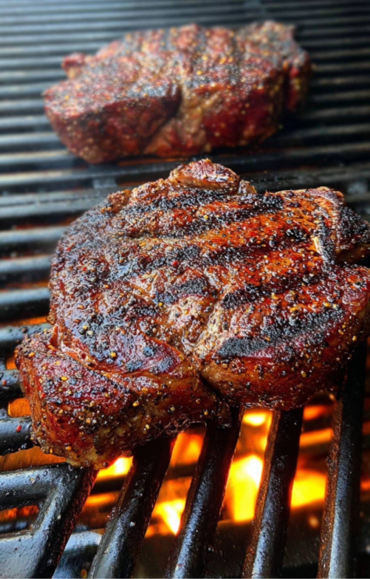 A close-up view of a ribeye steak on the grill showing a deep, caramelized brown crust and rendering fat over open flames.