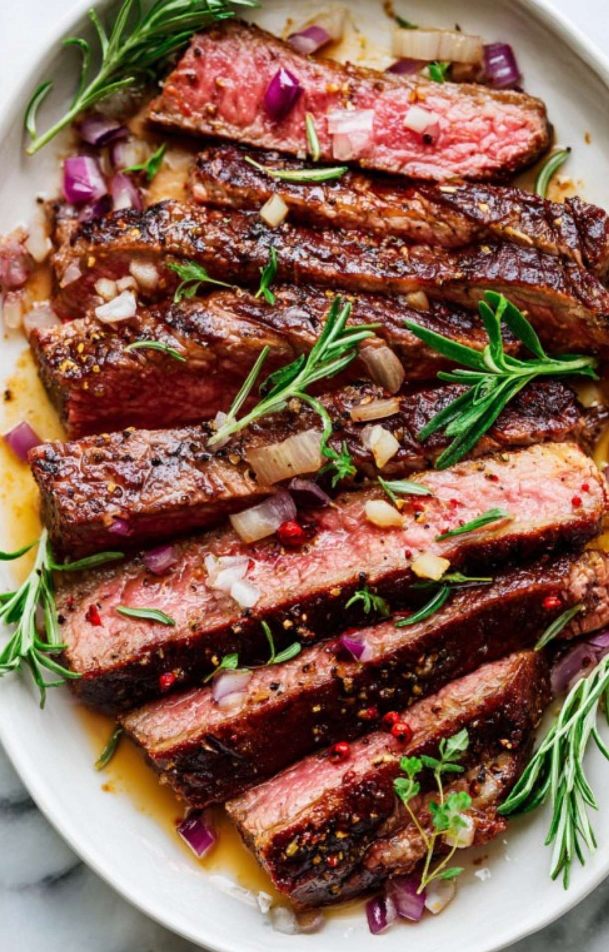 Sliced rib eye steak arranged on a white platter, topped with diced red onions, fresh herbs, and savory pan juices.