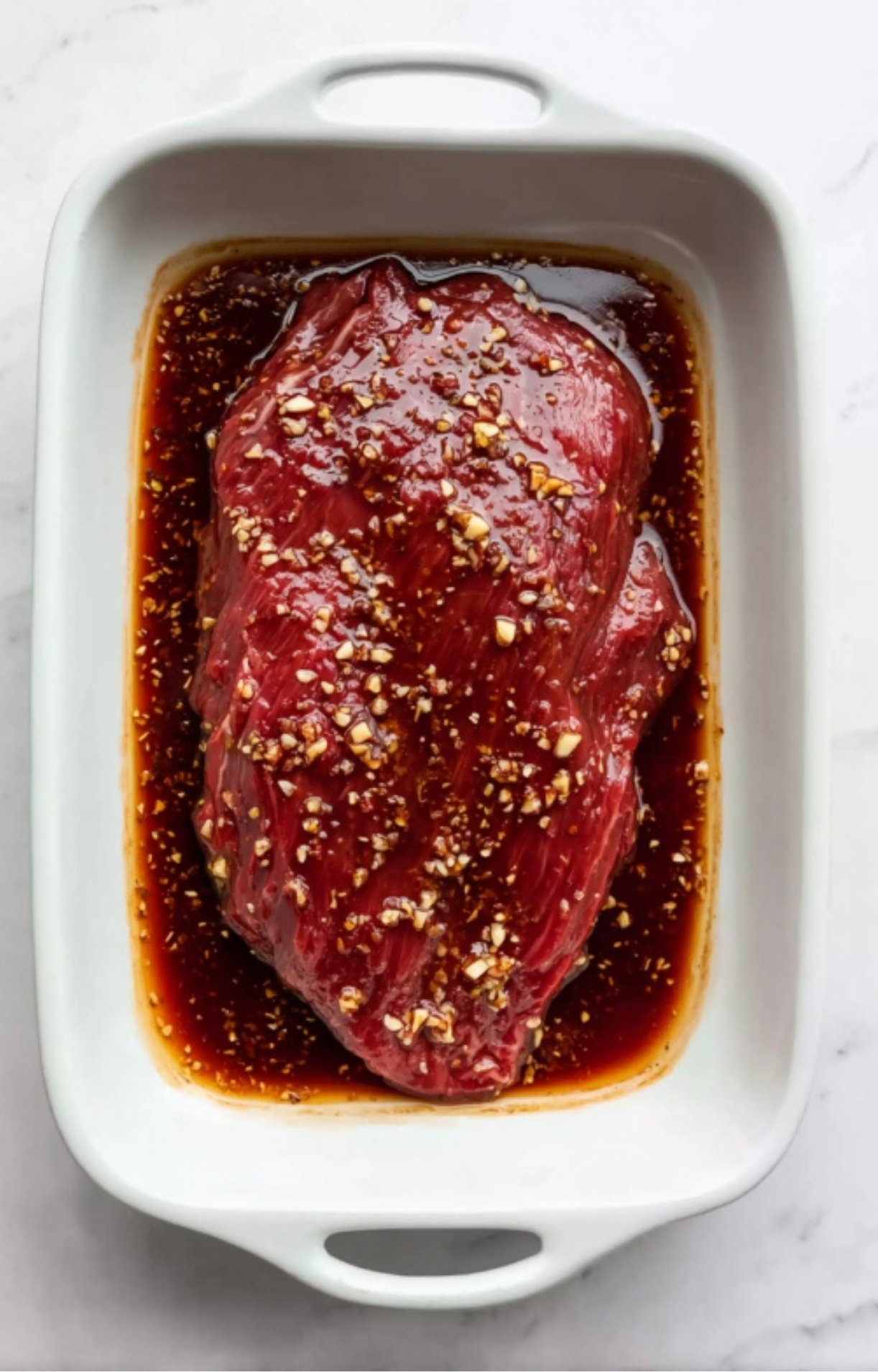 Raw flank steak soaking in a flavorful marinade inside a white baking dish.