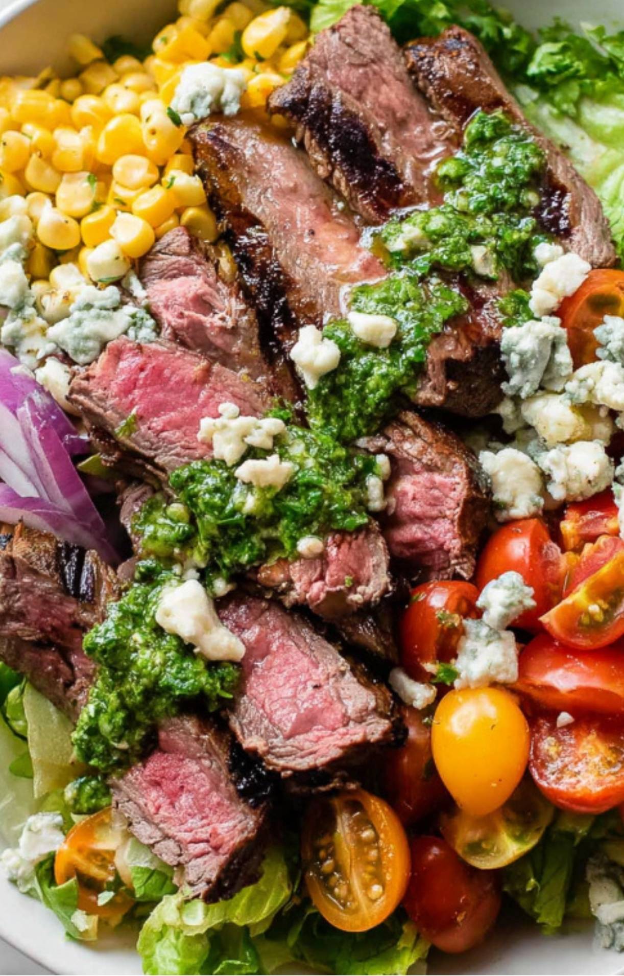 Close-up of sliced grilled steak on salad with chimichurri, blue cheese crumbles, corn, tomatoes, and lettuce, bright and colorful.