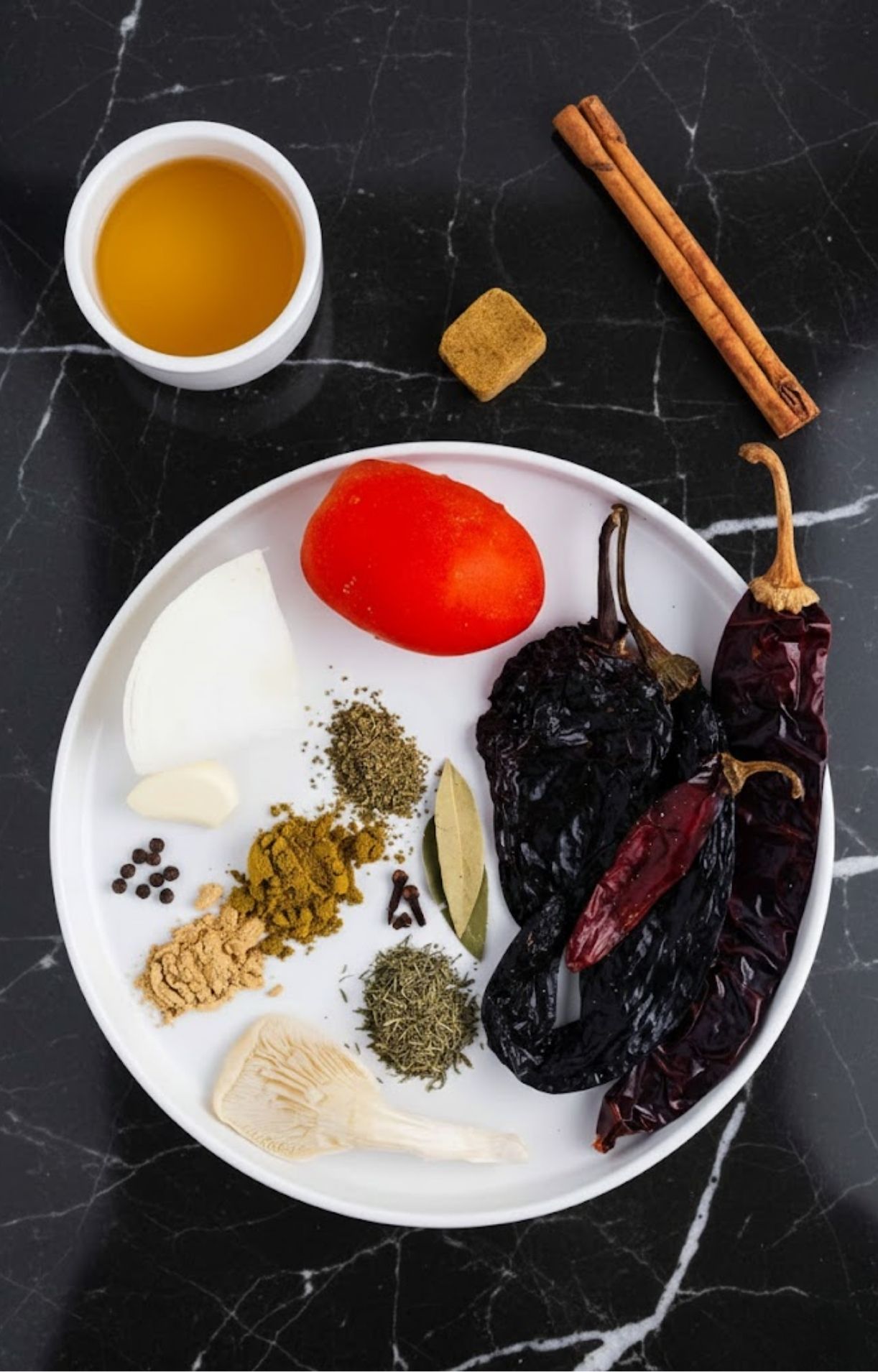 A flat lay of taco ingredients including dried chilies, a tomato, onion, garlic, spices (ginger, cumin, oregano, cloves, peppercorns), a bay leaf, cinnamon sticks, broth, and an oyster mushroom.
