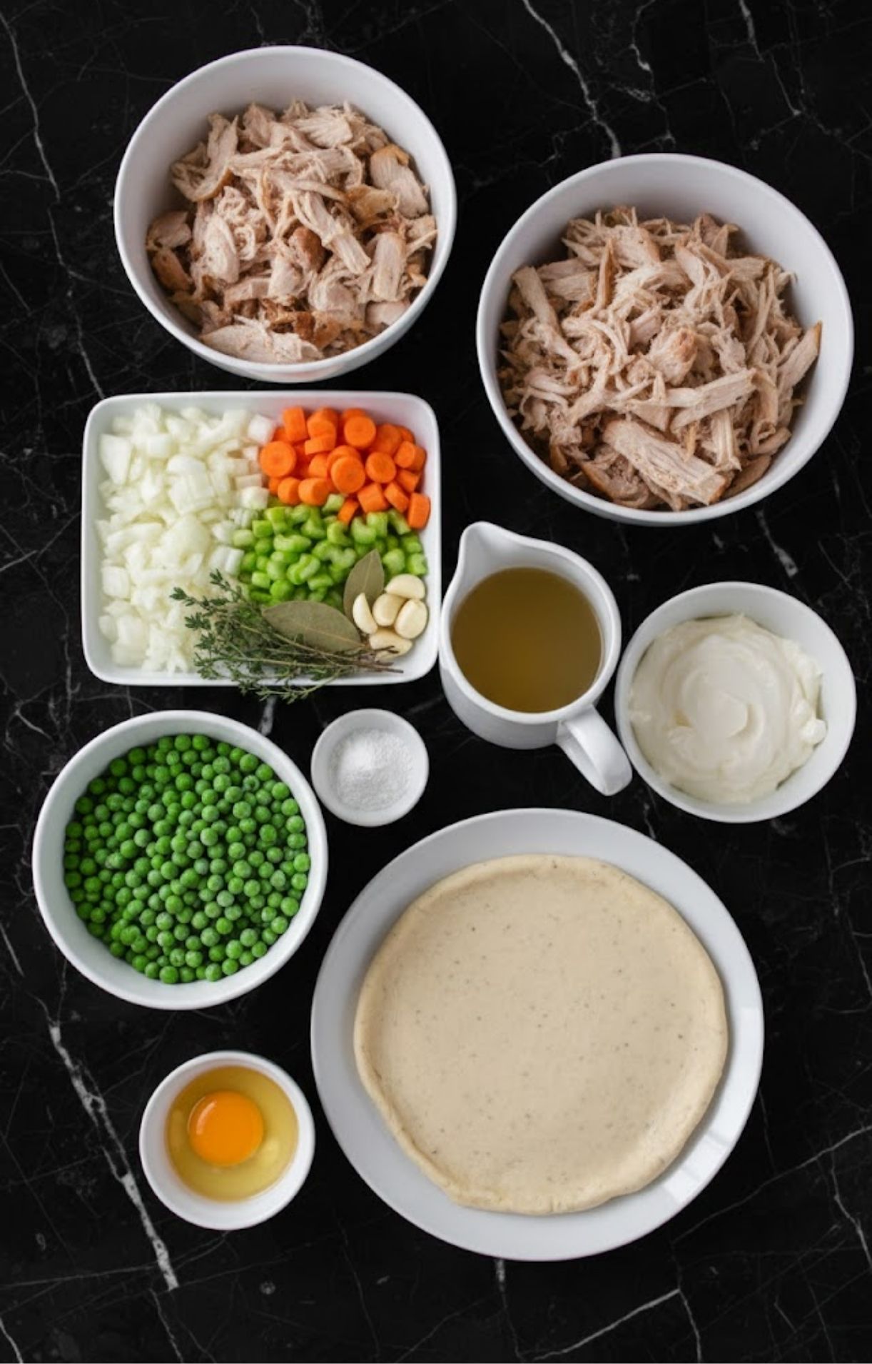 A flat-lay arrangement of bowls containing shredded turkey, peas, diced carrots, celery, onions, garlic, herbs, broth, cream, an egg, and a gluten-free pie crust on a black marble surface.