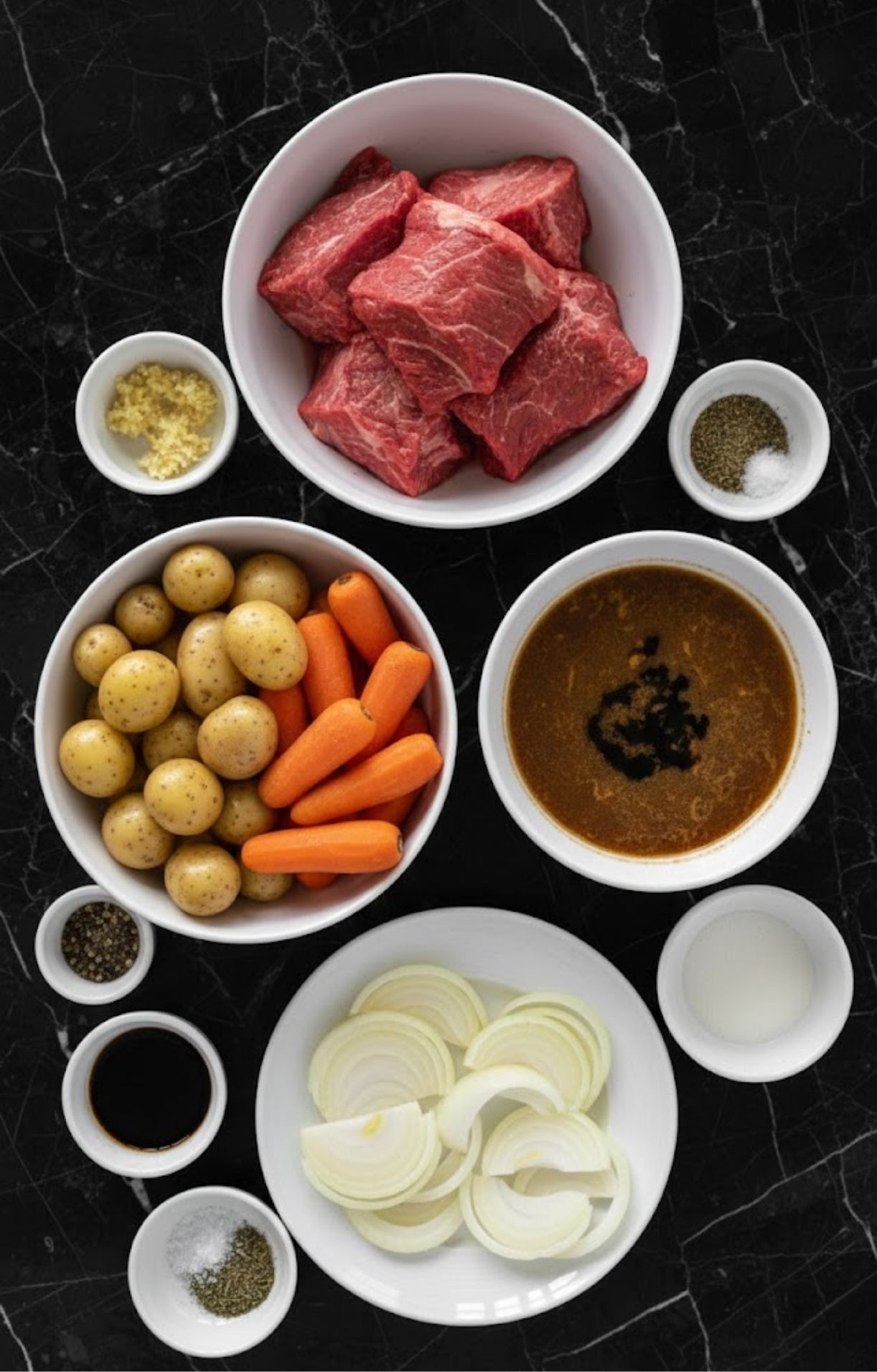 A top-down view of bowls containing raw chuck roast, baby potatoes, carrots, sliced onions, minced garlic, beef broth, and various seasonings on a black marble surface.