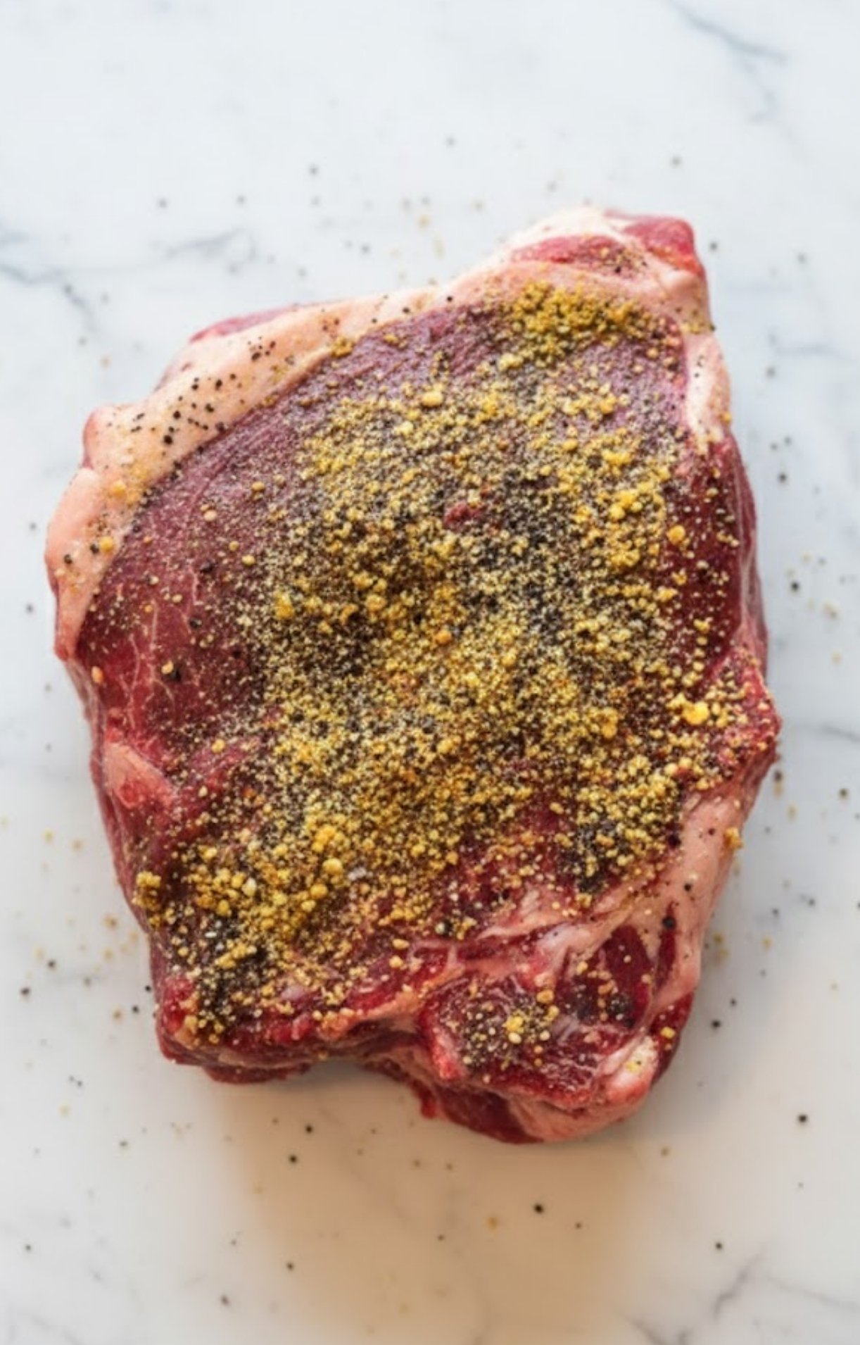 A raw beef chuck roast centered on a marble surface, generously coated with a yellow and black pepper dry rub seasoning.
