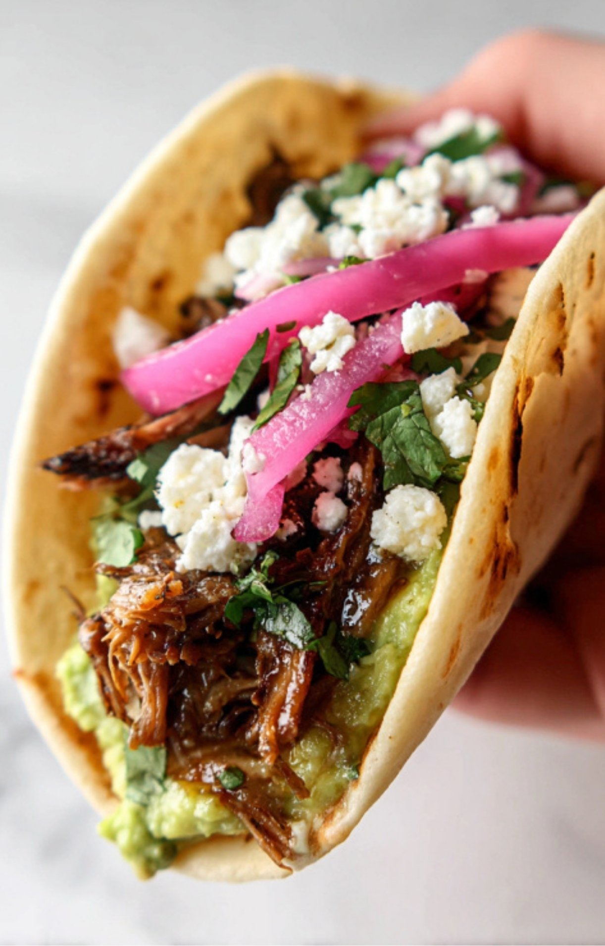 A close-up shot of a person holding a single taco filled with shredded chuck roast, creamy avocado mash, pickled red onions, and crumbled cotija cheese.