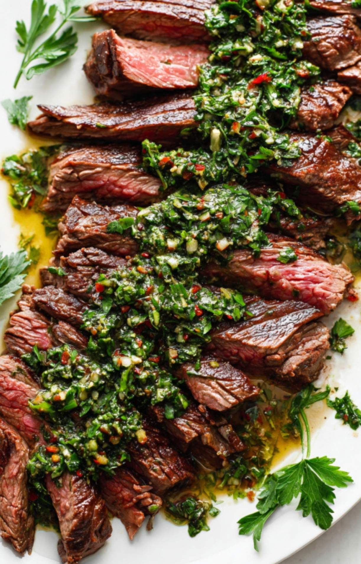 Sliced grilled skirt steak topped with vibrant chimichurri sauce, served on a white plate with fresh parsley leaves for garnish.