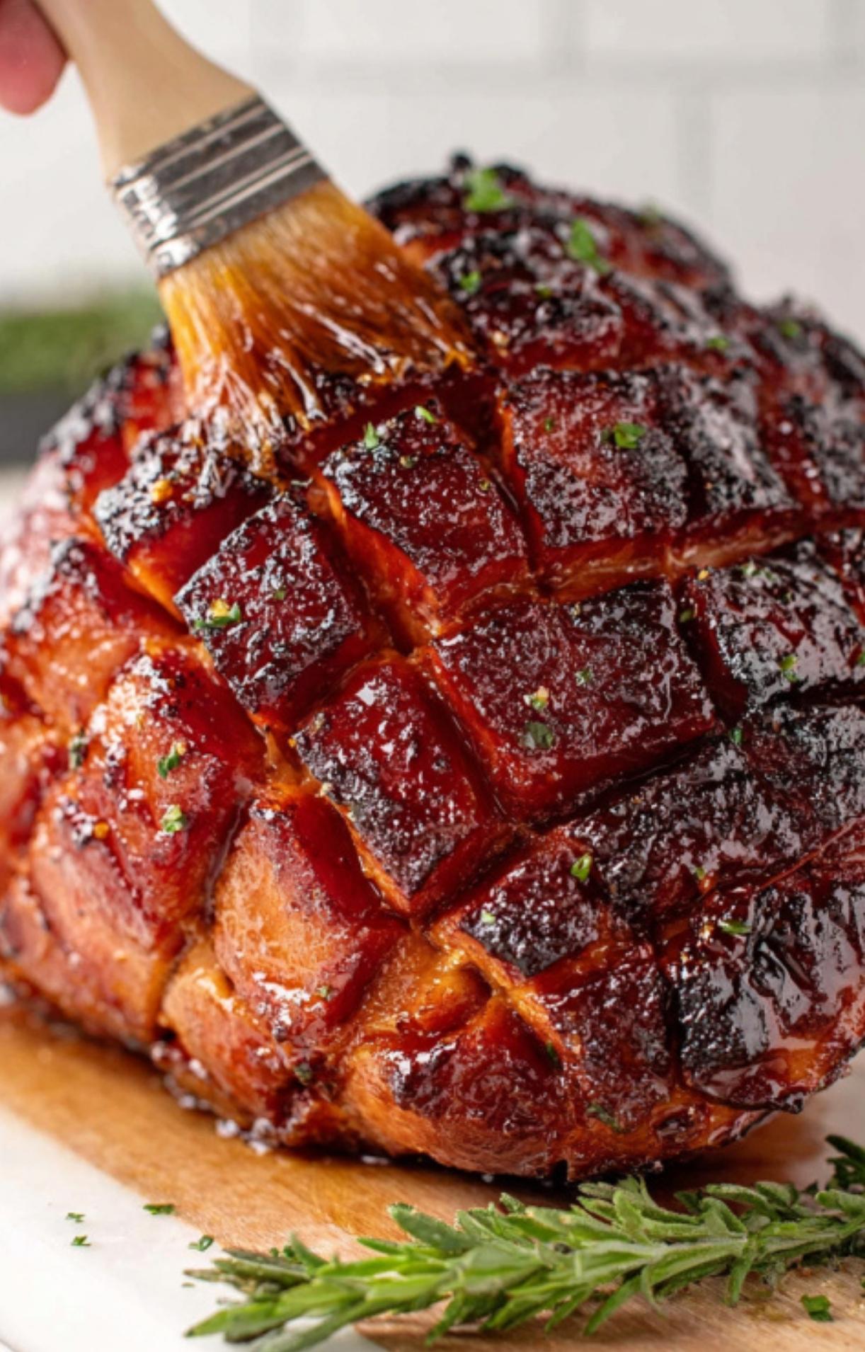 A hand using a pastry brush to add sweet cherry glaze to a hot, caramelized ham for extra flavor and shine.