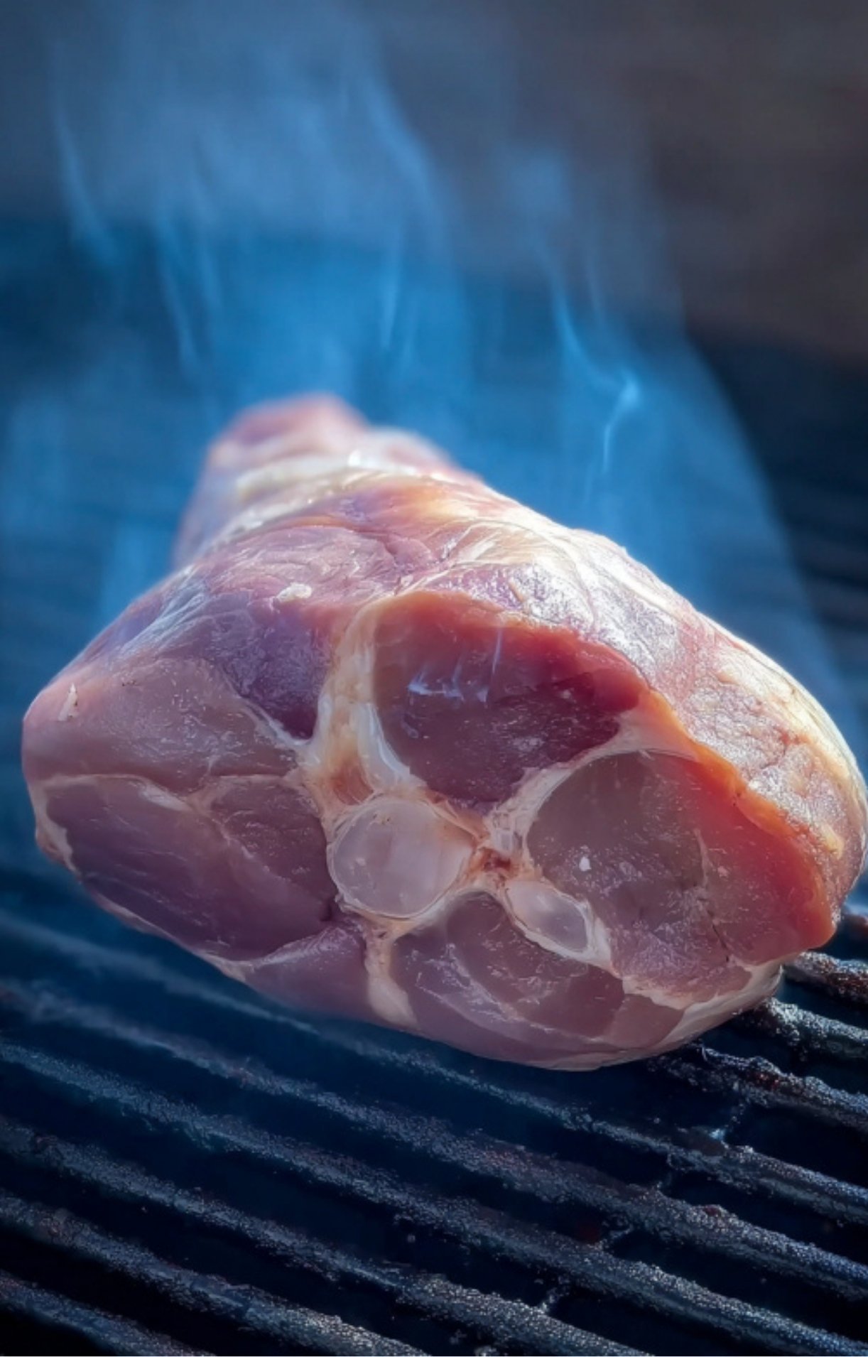 A raw, cross-cut ham hock sits on a black grill grate as thin wisps of blue smoke rise around the pink meat.