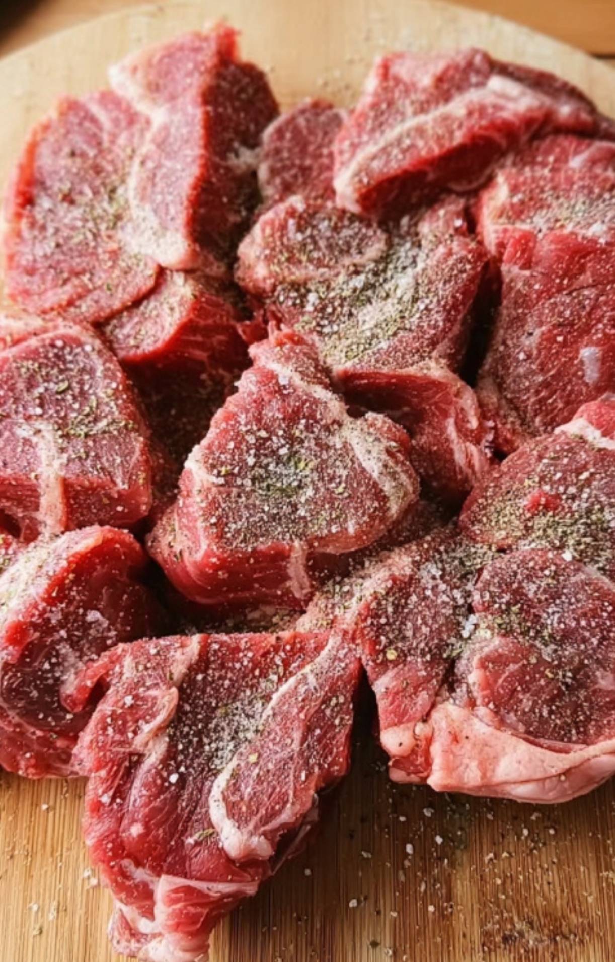 Raw chunks of beef chuck roast spread out on a wooden cutting board and generously seasoned with salt, pepper, and dried herbs.