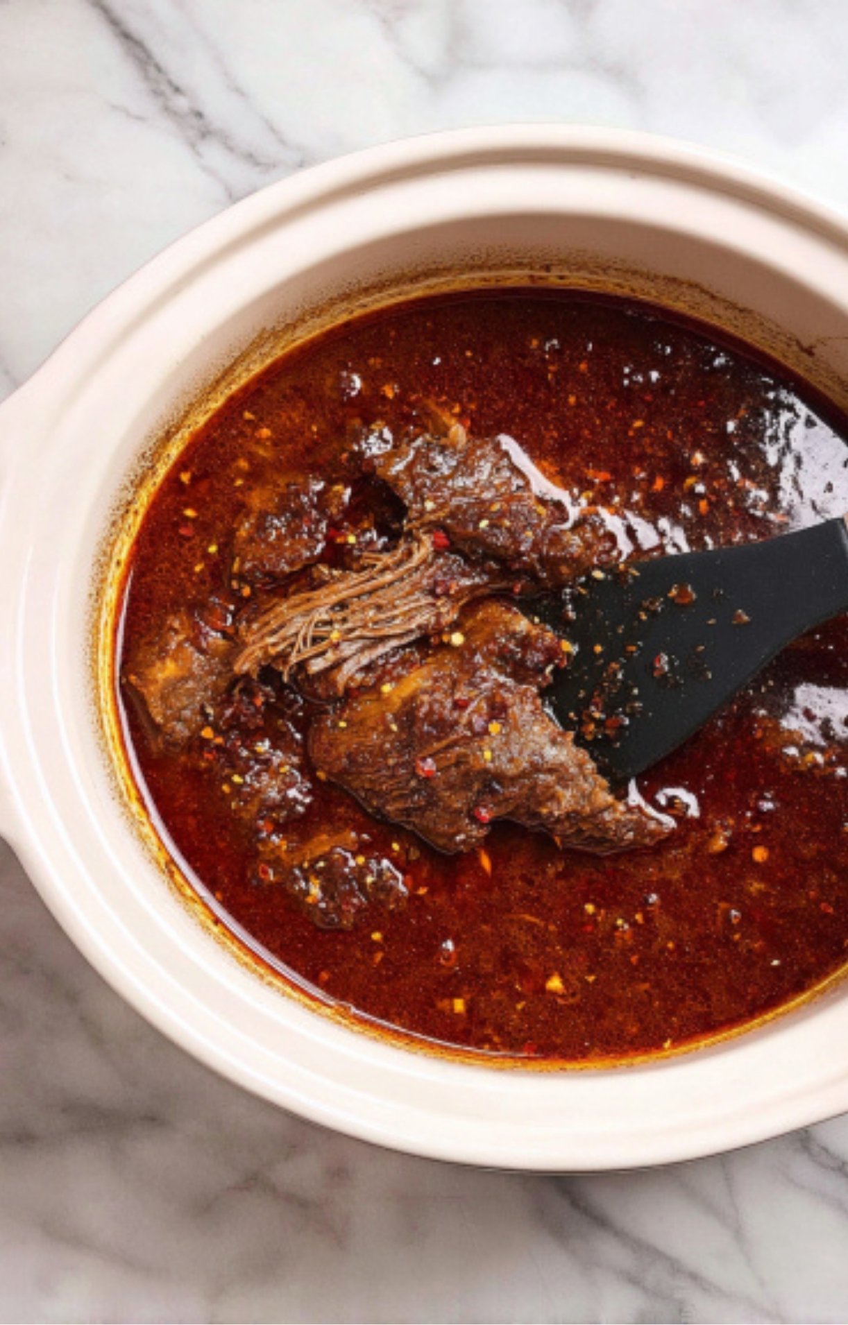 Large chunks of beef simmering in a deep red, spiced birria broth inside a white ceramic slow cooker or Dutch oven.