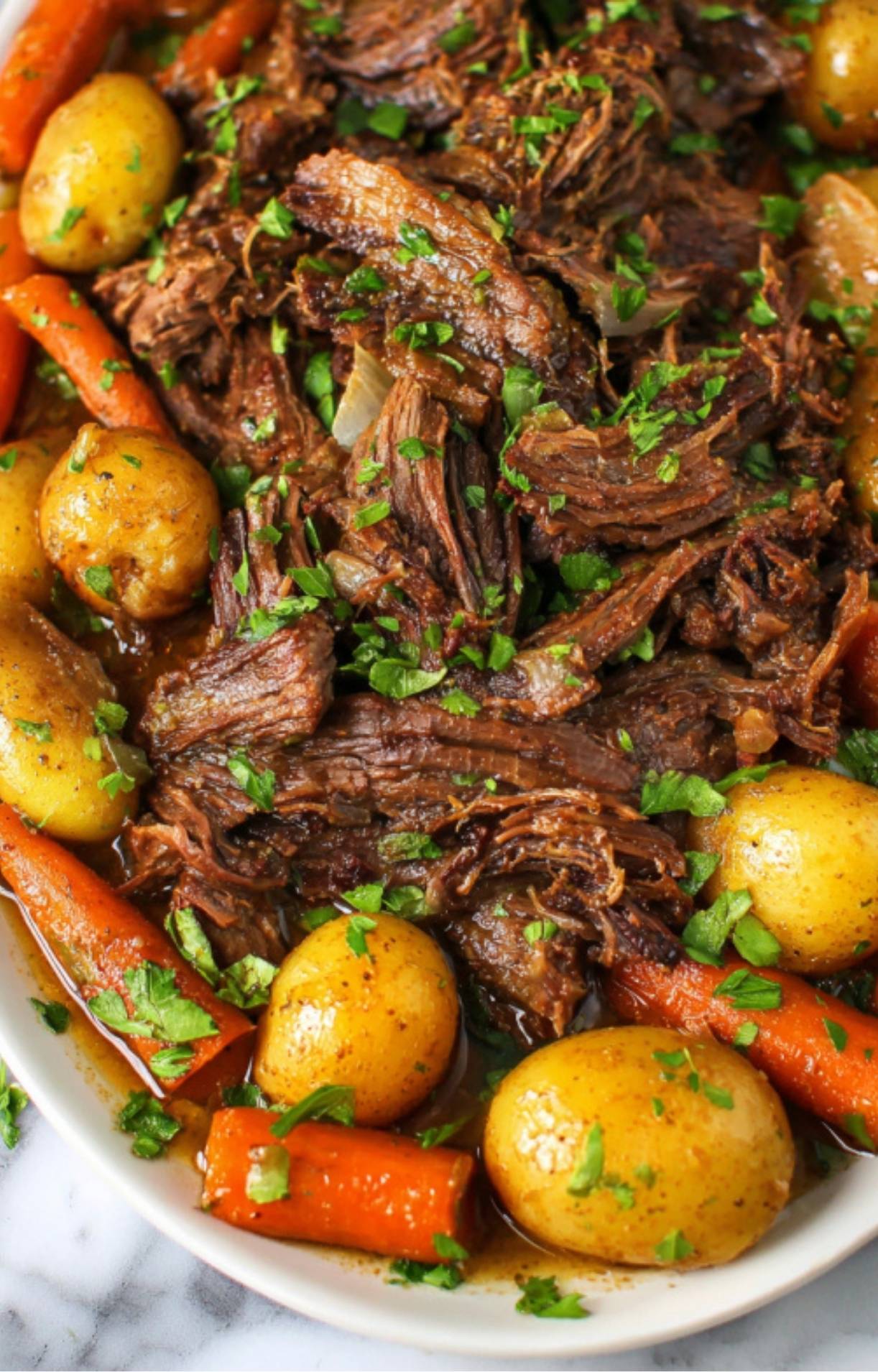 A white plate featuring a serving of shredded beef pot roast alongside cooked baby potatoes and carrots, garnished with fresh parsley.