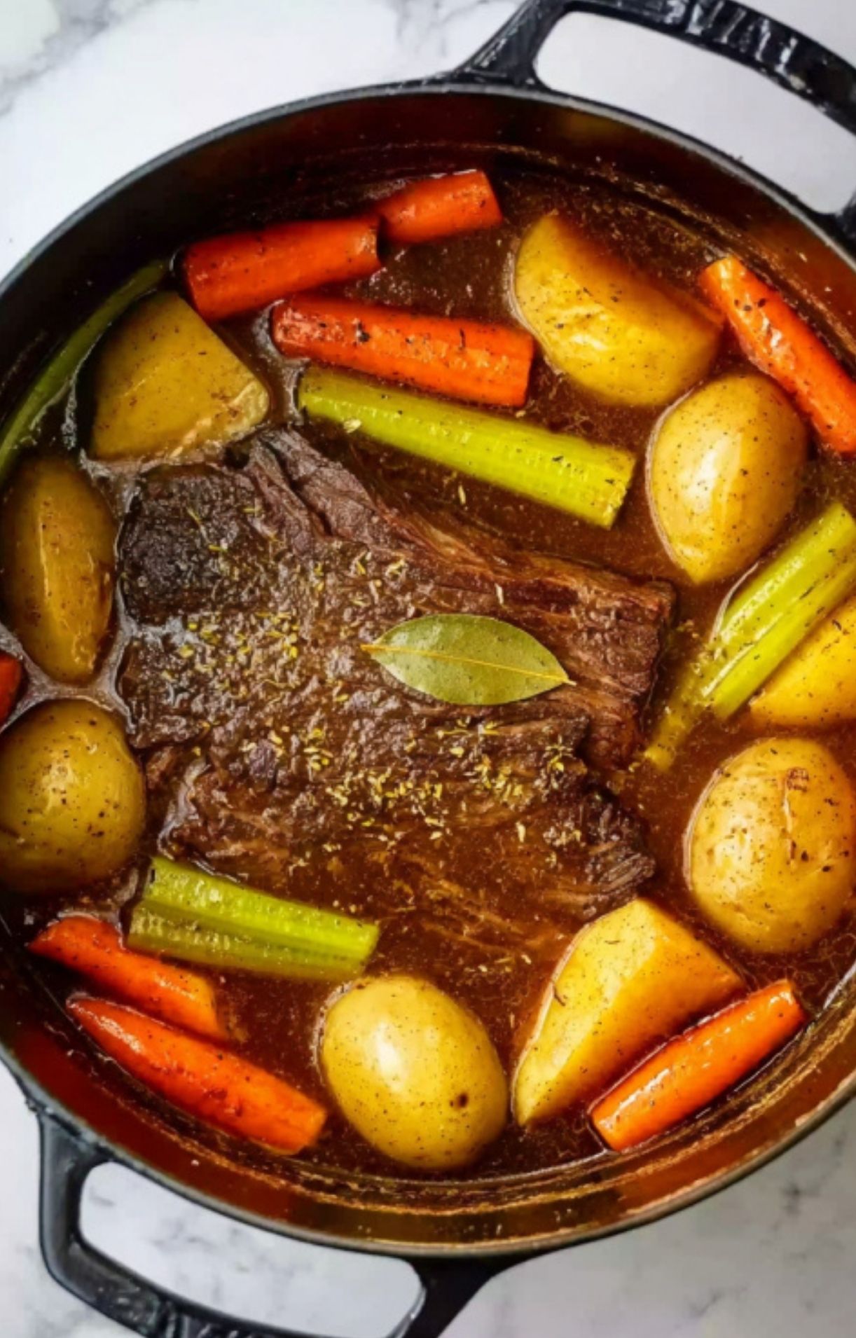 The seared roast in the Dutch oven surrounded by potatoes, carrots, celery, and a bay leaf in a dark savory liquid.