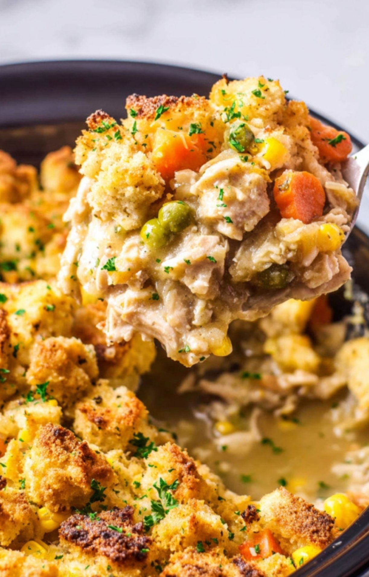 A large metal spoon lifting a hearty portion of turkey, carrots, and peas topped with a crispy, buttery stuffing crust.