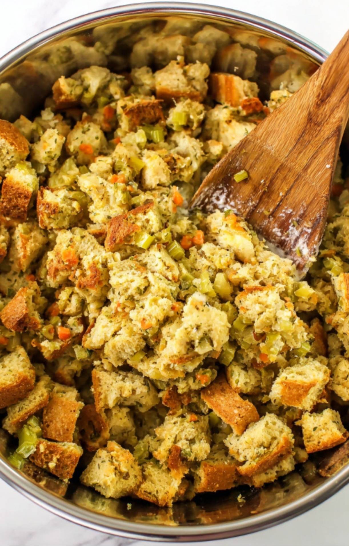 A wooden spoon mixing cubes of bread stuffing with herbs and celery in a metal bowl.