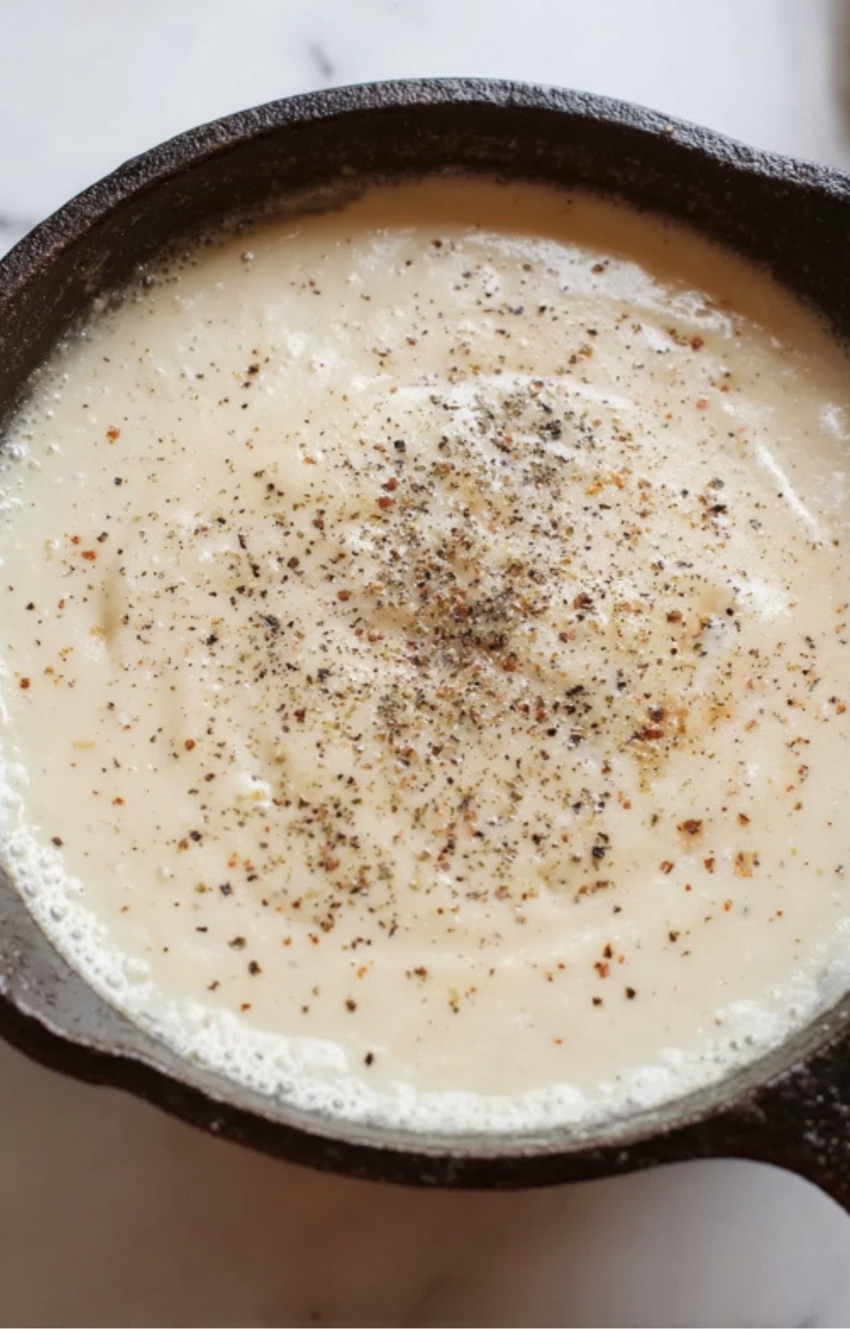 A creamy white sauce in a skillet topped with a generous sprinkle of black pepper and herbs.