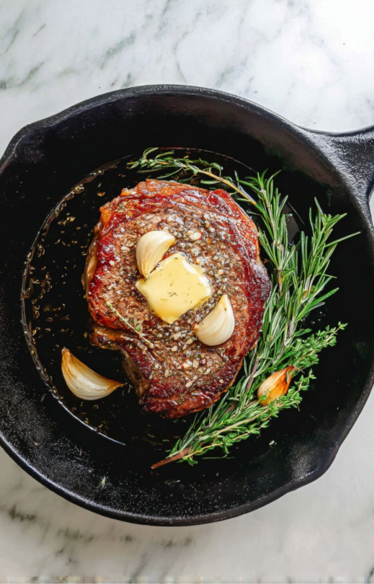 A seared rib eye steak in a skillet topped with a pat of butter and garlic cloves, surrounded by fresh sprigs of rosemary and thyme.