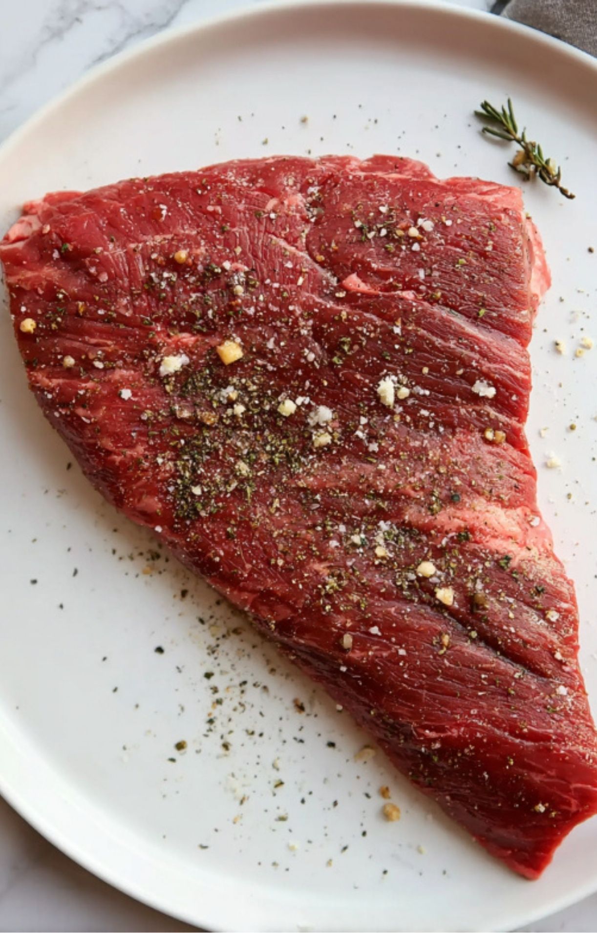 Raw tri tip steak sprinkled with salt, pepper, and spices on a white plate, prepped for grilling.