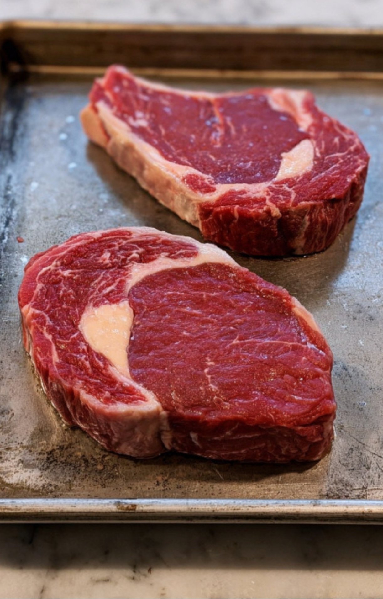 Two fresh, thick-cut raw rib eye steaks with beautiful marbling resting on a metal baking tray.