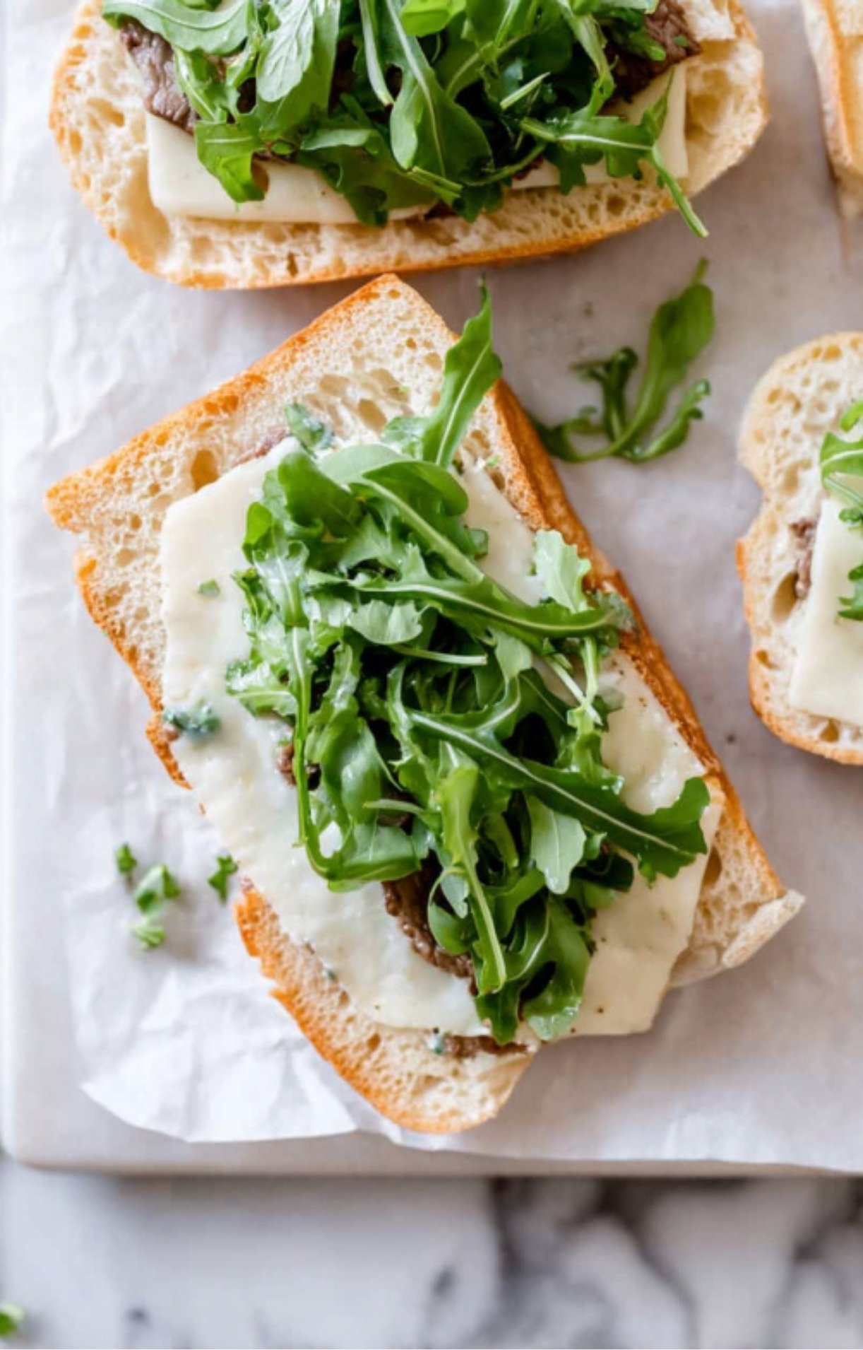 Open-faced ciabatta bread topped with melted white cheese and a handful of fresh green arugula.