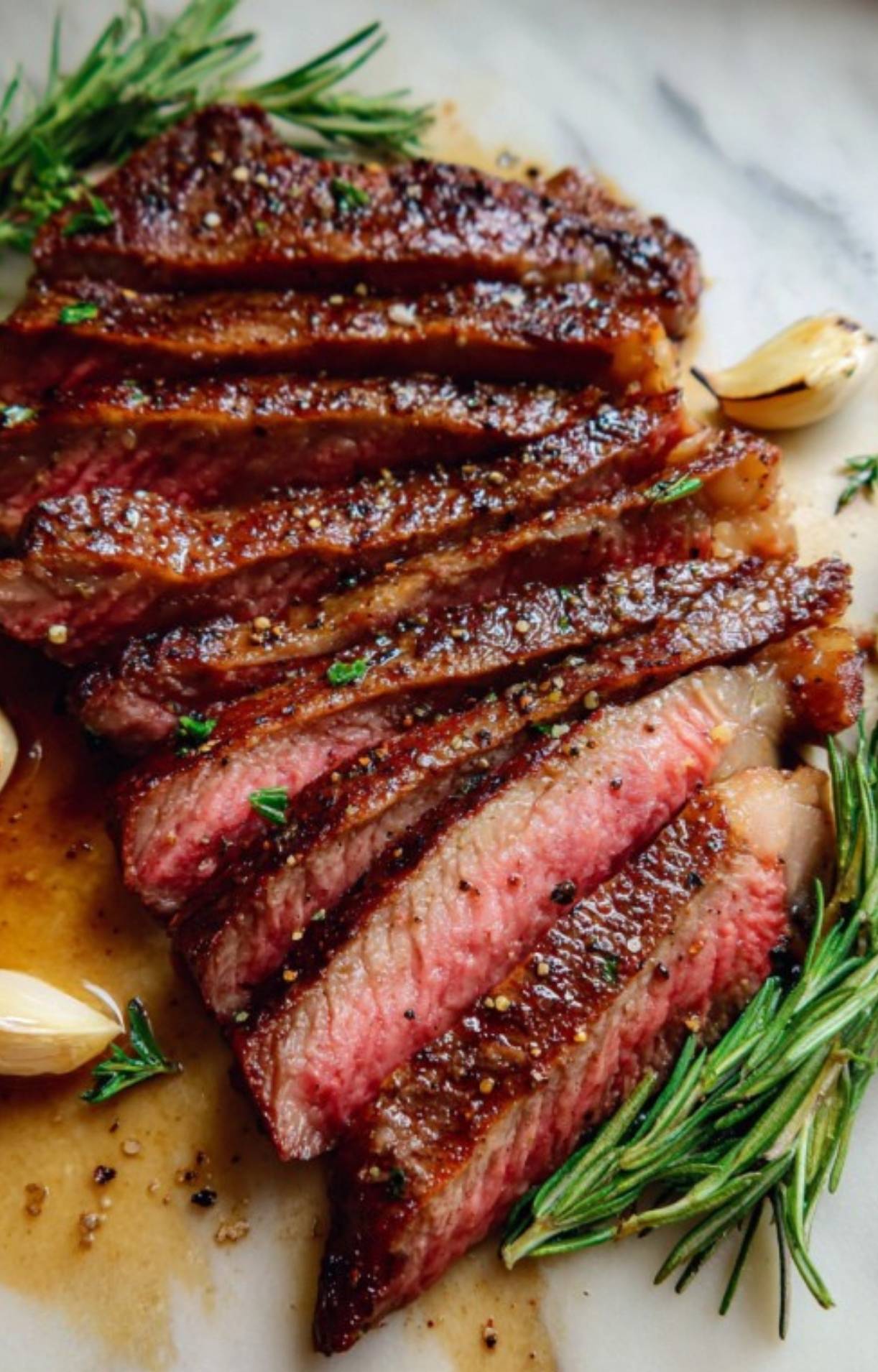 Close-up of thin sliced rib eye steak showing a perfect medium-rare center, garnished with garlic cloves and fresh rosemary.