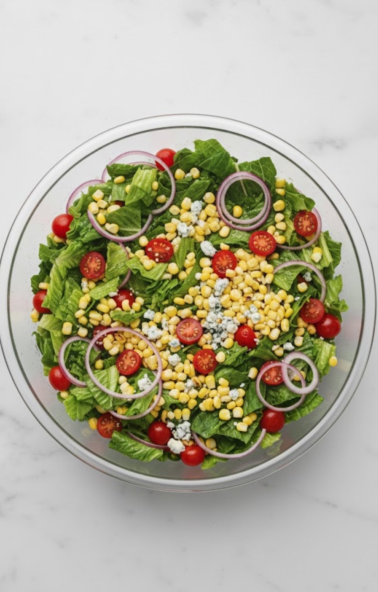 Large glass bowl filled with chopped romaine, cherry tomatoes, corn, red onion rings, and blue cheese ready to toss for a salad.