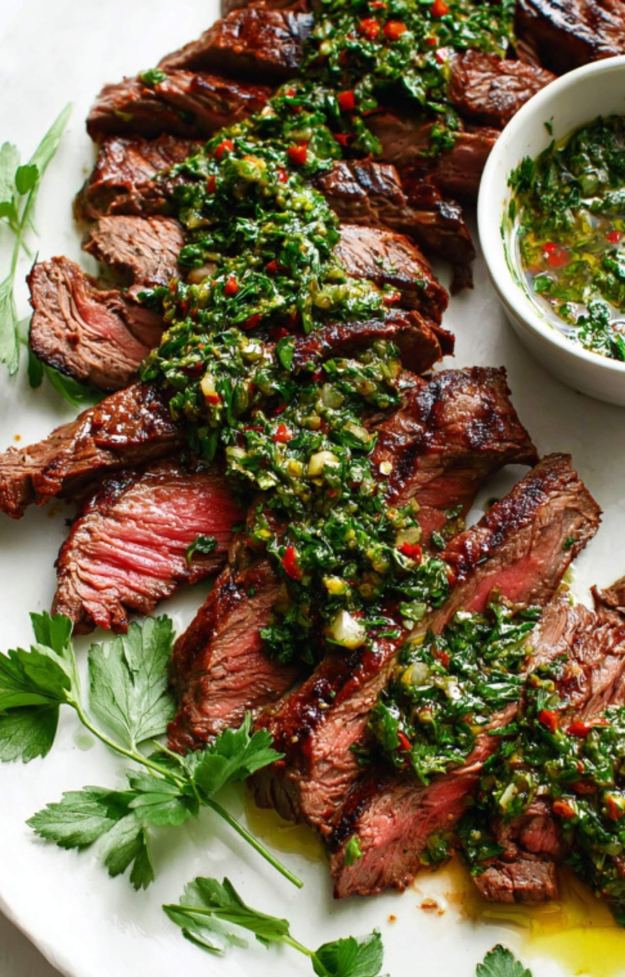 Juicy grilled skirt steak slices arranged on a platter, generously topped with homemade chimichurri sauce and garnished with parsley, with a small bowl of extra sauce on the side.