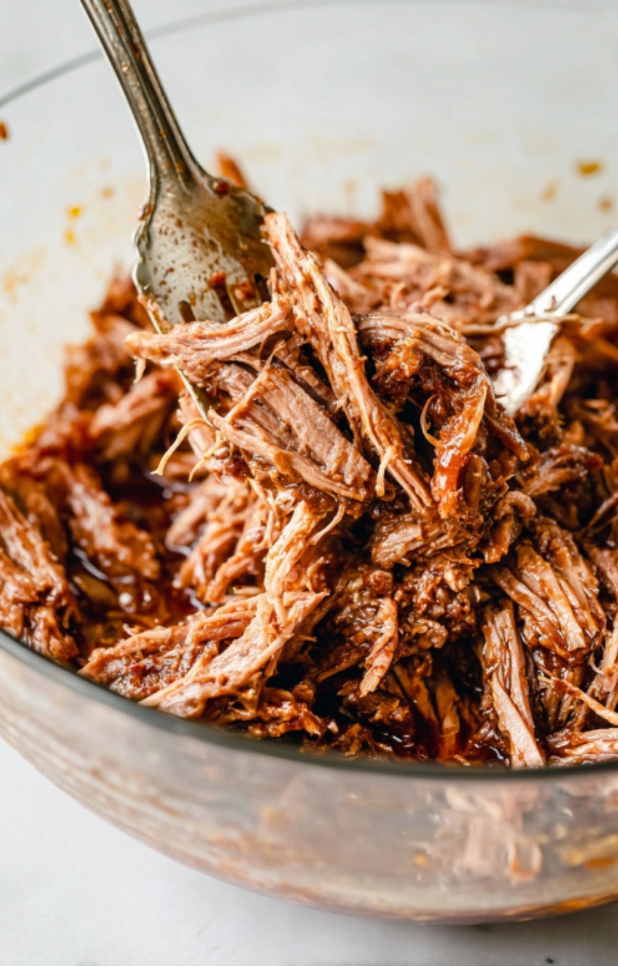 Two silver forks shredding tender, juicy braised beef in a glass bowl to prepare the filling for Crockpot Birria Tacos.