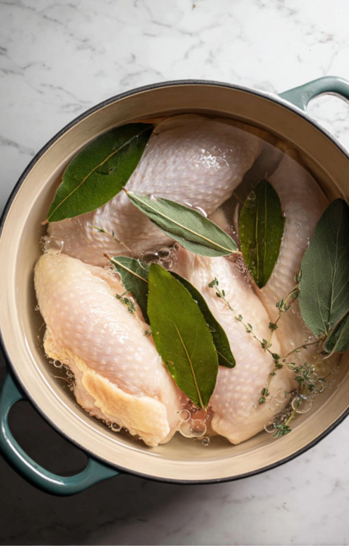 Chicken breasts simmering in a pot of water with fresh bay leaves and thyme.