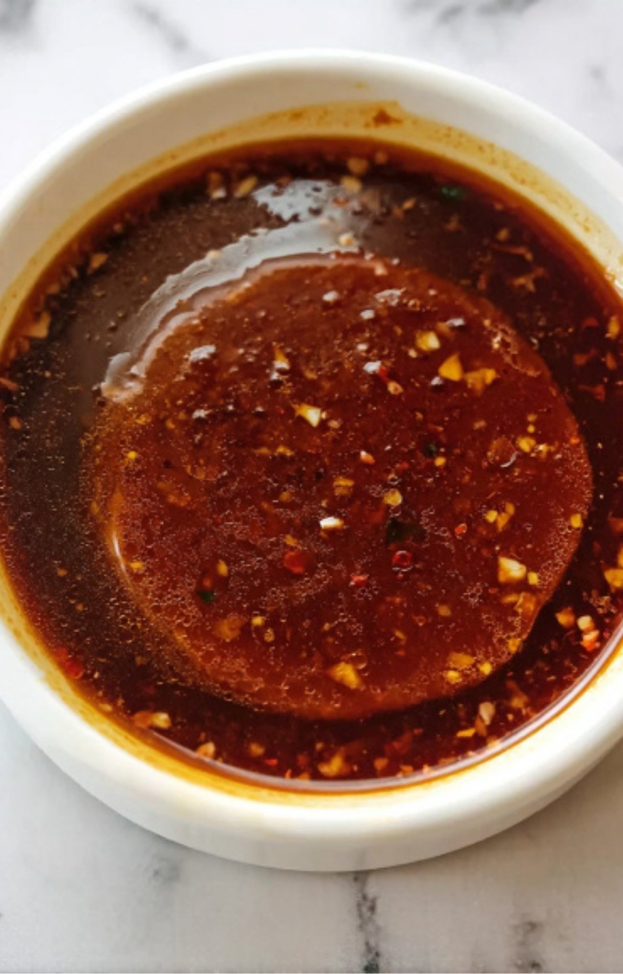 A top-down view of a bowl filled with dark red birria broth, showing droplets of flavorful fat and bits of minced garlic on the surface.