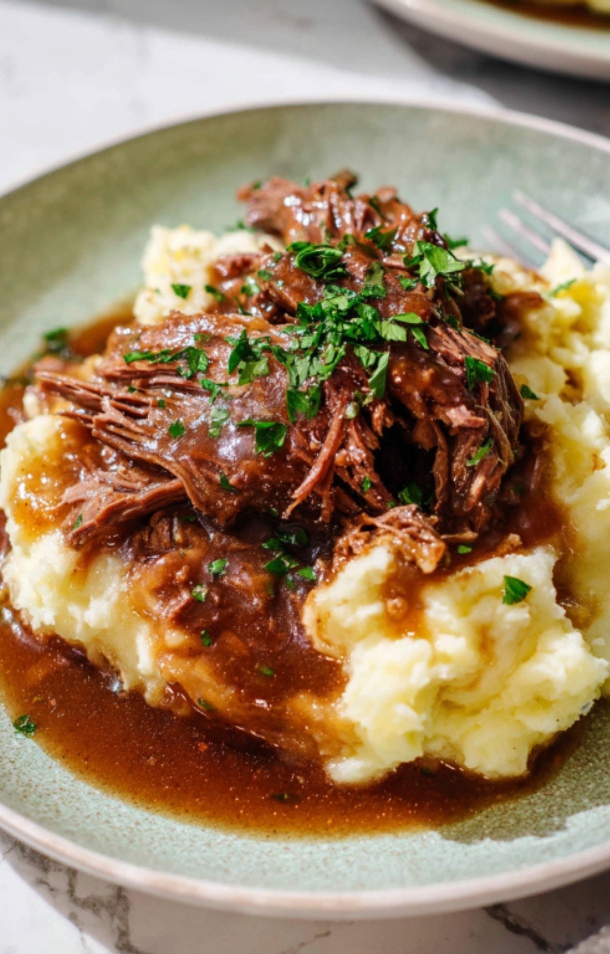 A close-up of tender, shredded braised beef served over a bed of fluffy mashed potatoes, smothered in a rich brown gravy and garnished with fresh chopped parsley.