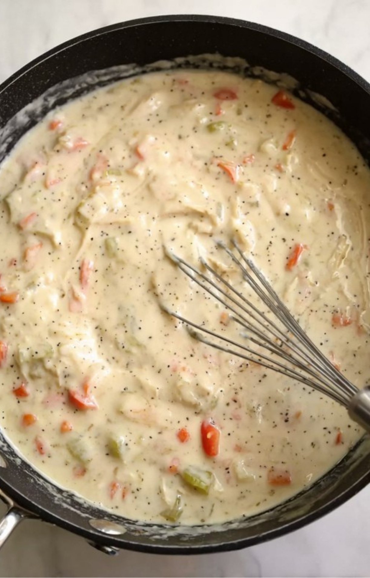 A wire whisk blending a smooth, peppery white cream sauce with vegetables in a large skillet.